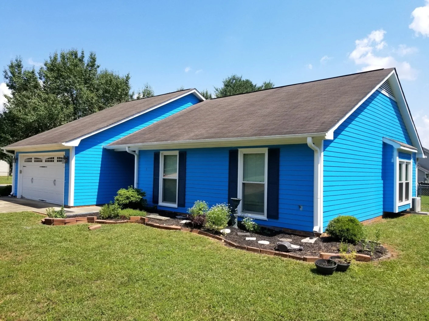 Bright blue house with dark shutters, a white garage door, neatly landscaped yard with shrubs, flowers, and a garden bed, under a partly cloudy sky.
