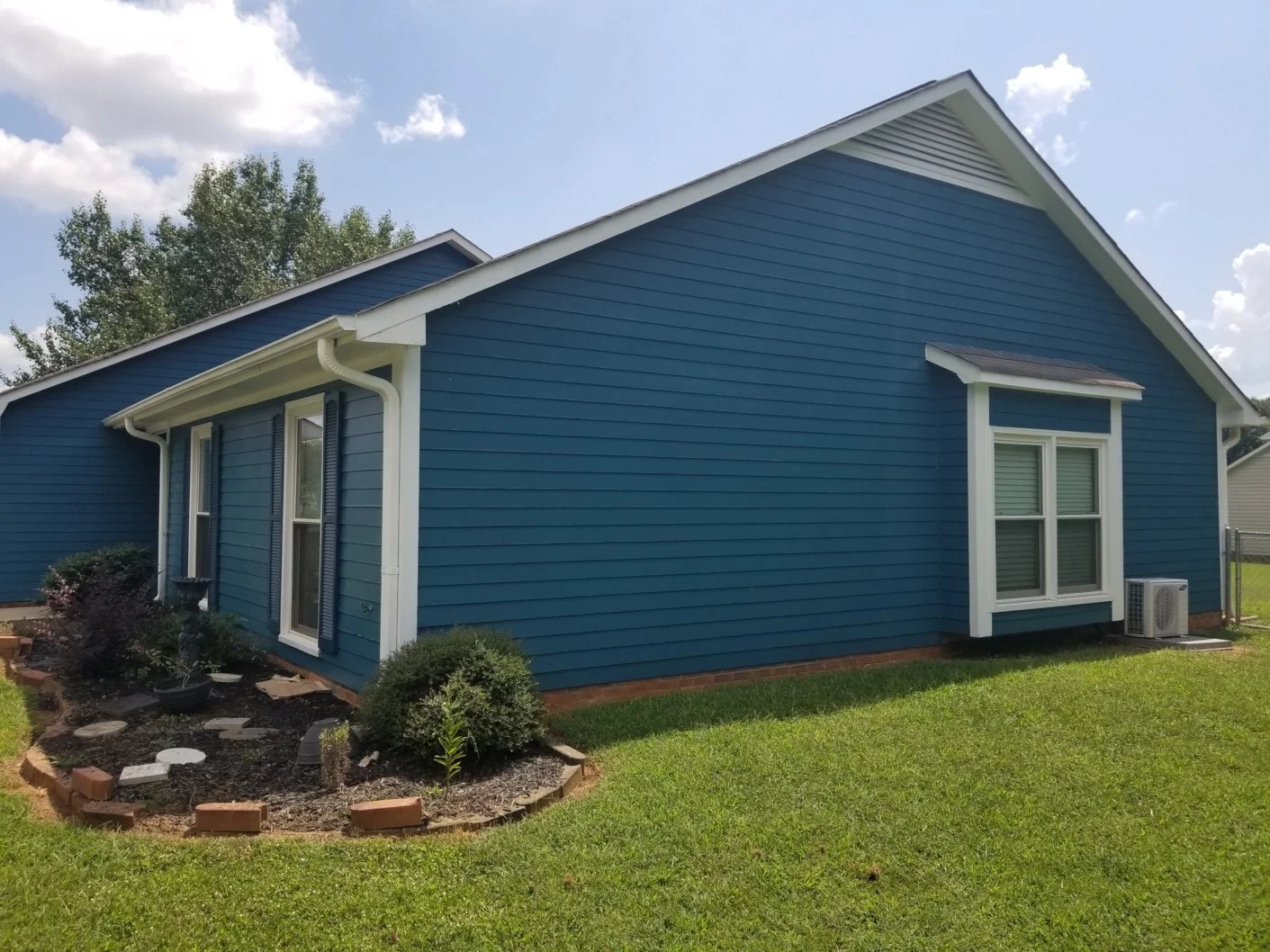 A blue house with white trim, two windows, green lawn, and a small garden bed with bushes and stepping stones.