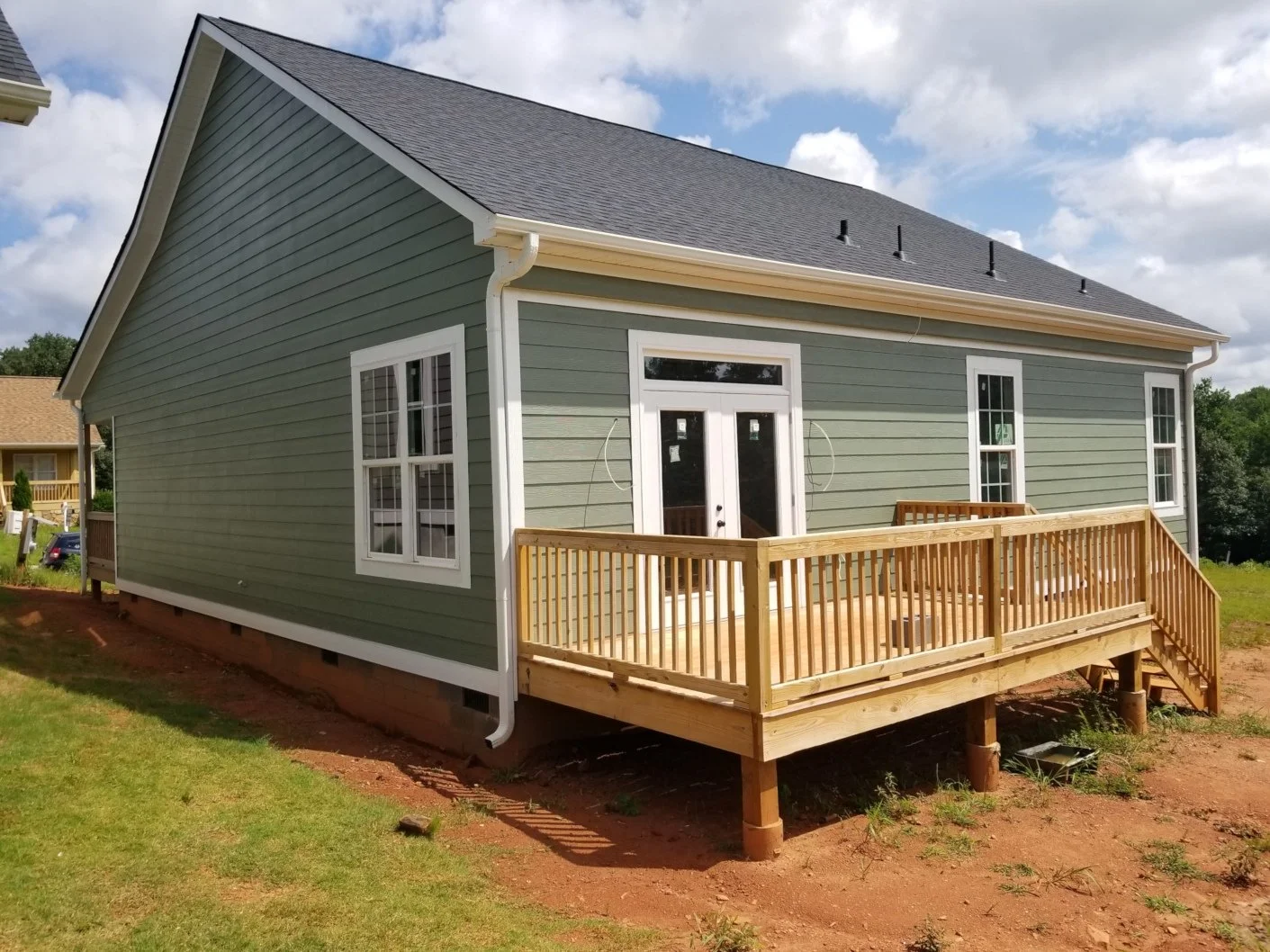A newly built house with green siding, white trim, and a dark gray roof. It features a small wooden deck with stairs and a railing, and multiple windows.