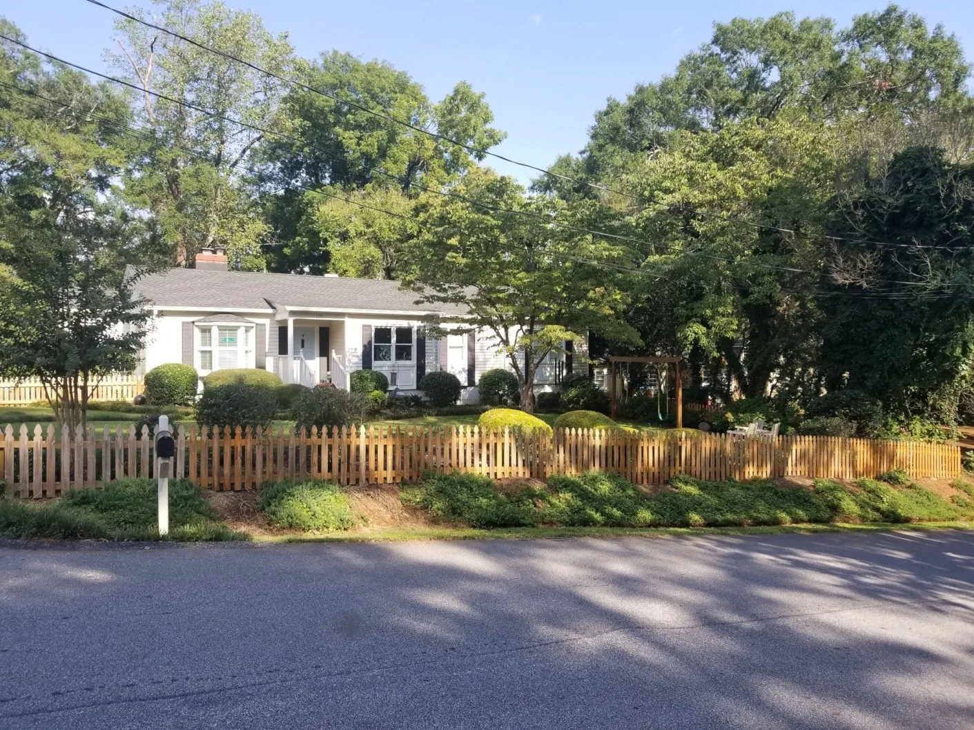 A house with a front yard, surrounded by trees, shrubbery, and a wooden fence, with a driveway in the foreground.
