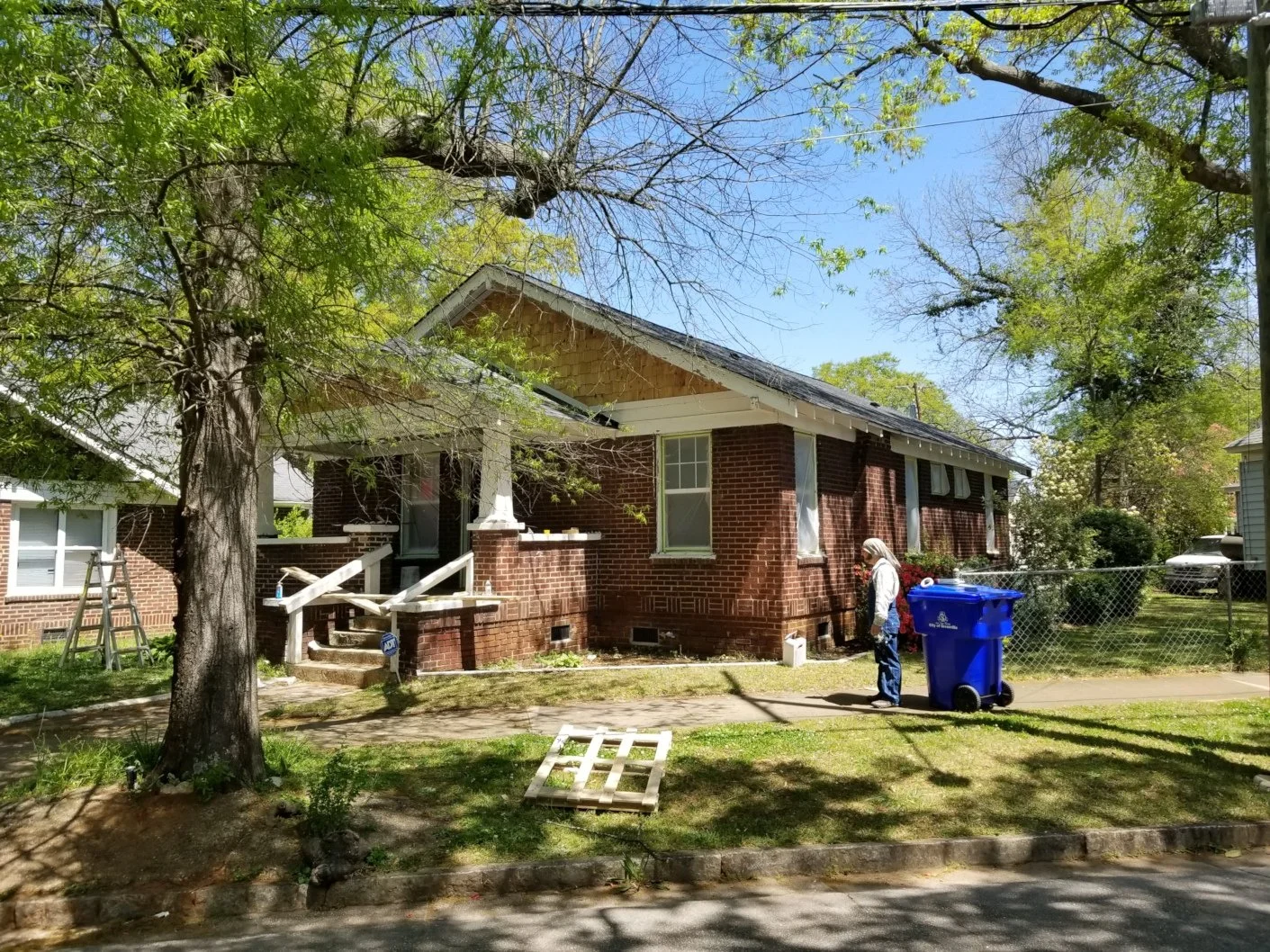 A person standing on sidewalk near blue trash bin in front of brick house with trees and blue sky