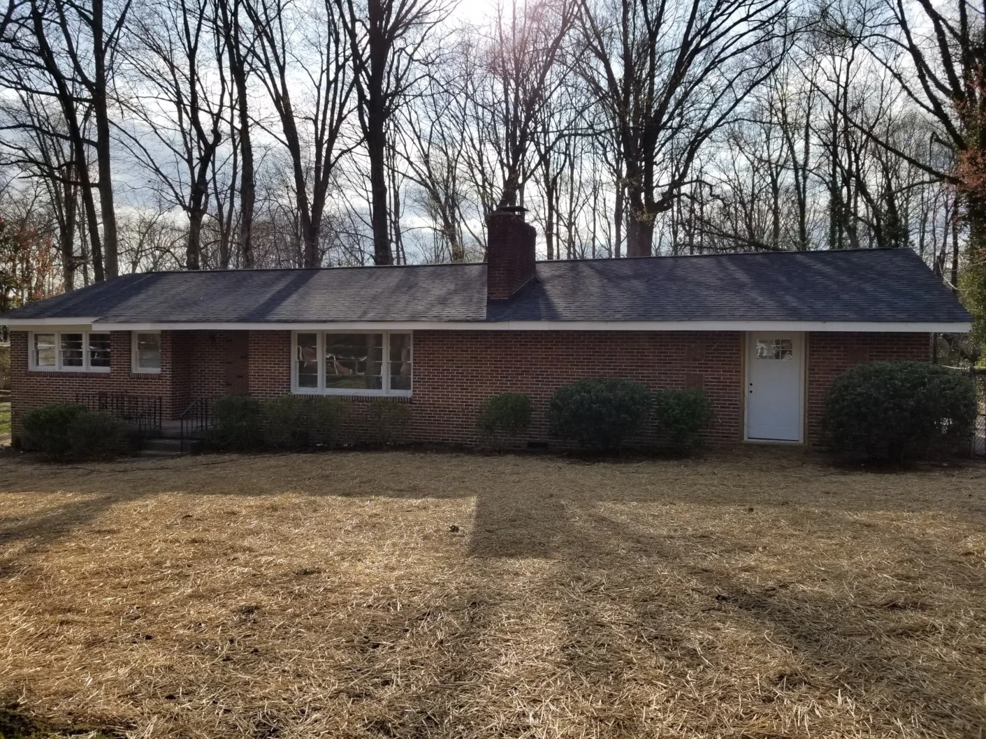 A brick house with a dark shingled roof, large front windows, a white front door, and bushes along the front. There are leafless trees in the background and a dry grassy yard in the foreground.
