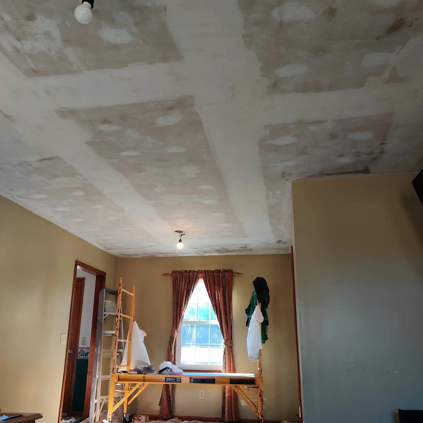 Interior room with a ceiling undergoing drywall repair, scaffolding with tools and clothes, a window with curtains, and a light bulb hanging from the ceiling.