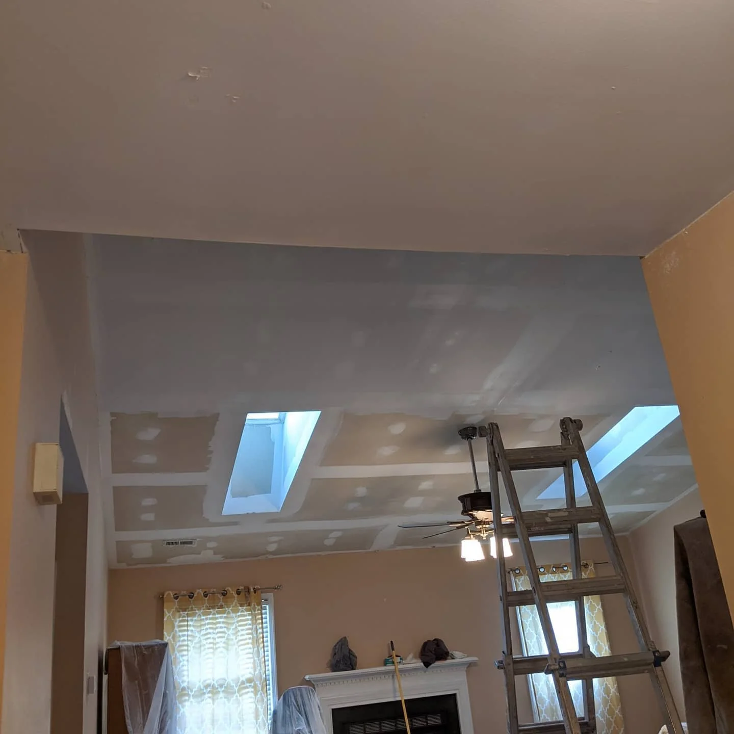 Living room ceiling under renovation with drywall patches, skylights, a ladder, ceiling fan, and window with yellow patterned curtains.