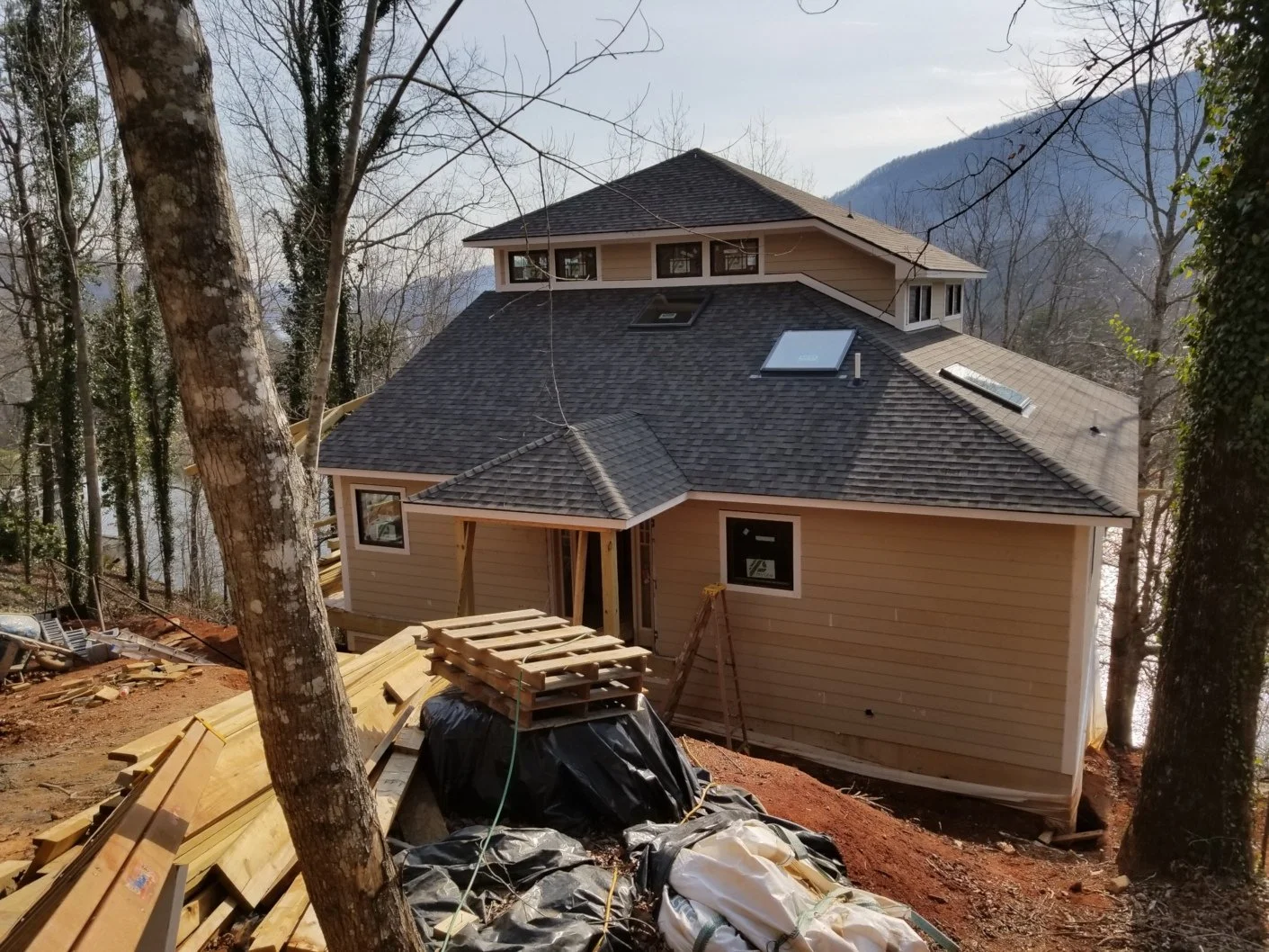 A house under construction on a hillside surrounded by trees, with mountains in the background. The house has a dark shingled roof, skylights, and beige siding. Construction materials and tools are scattered around the site.