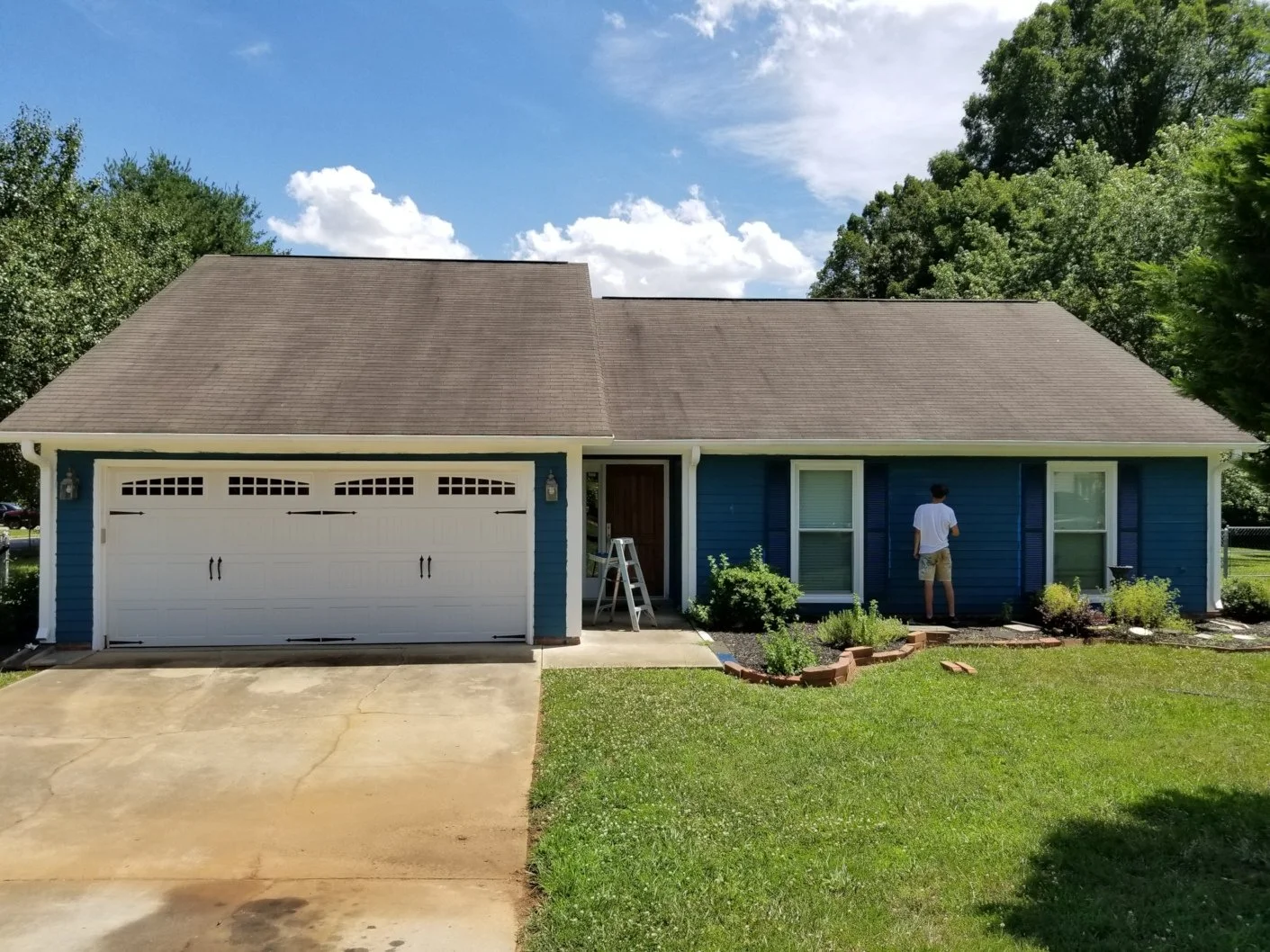 A blue house with a white garage door, front lawn, and a man standing near the windows on a bright sunny day.