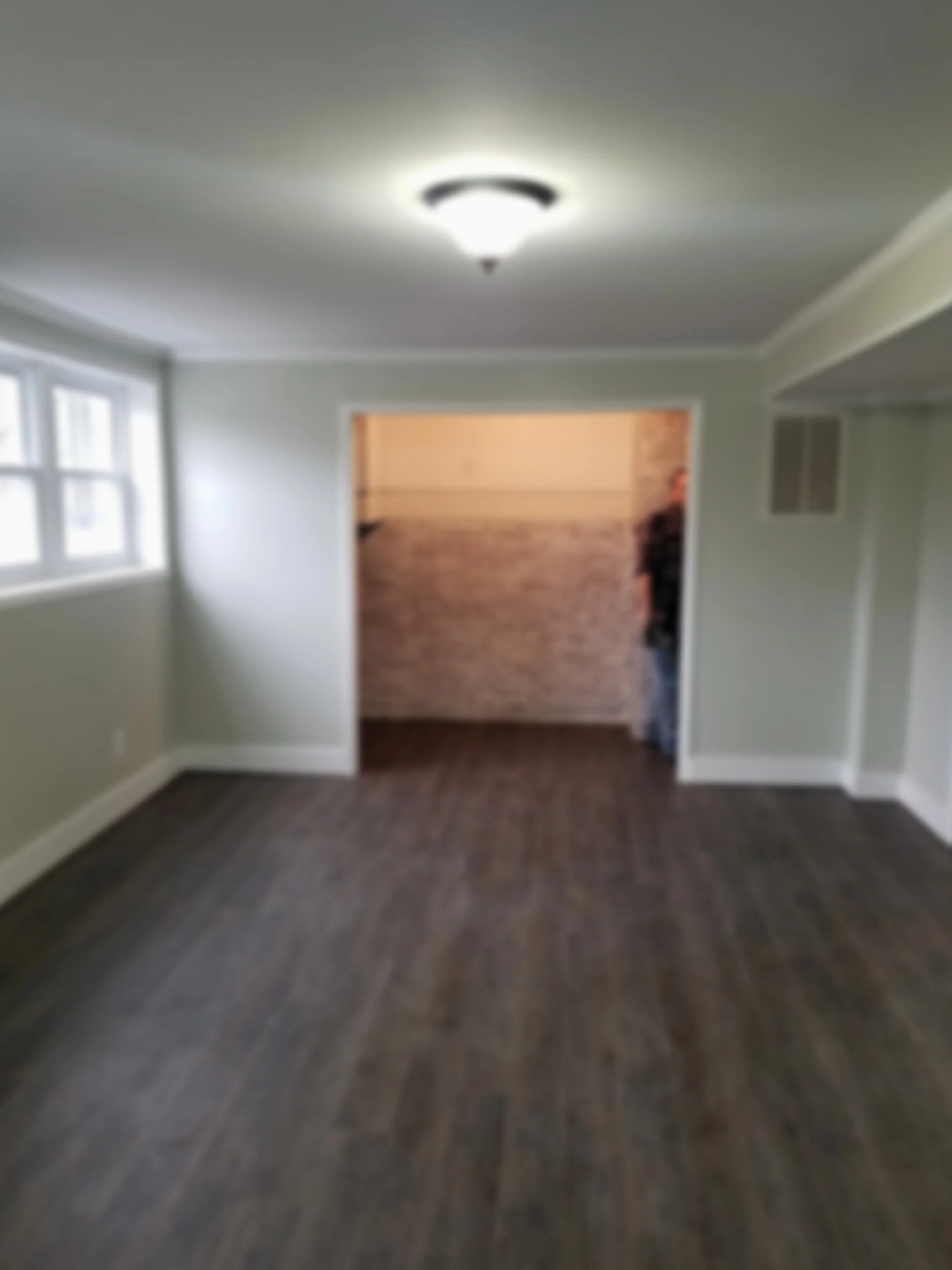 Empty room with hardwood floors, white walls, a window on the left, and a doorway leading to another room with a person partially visible.