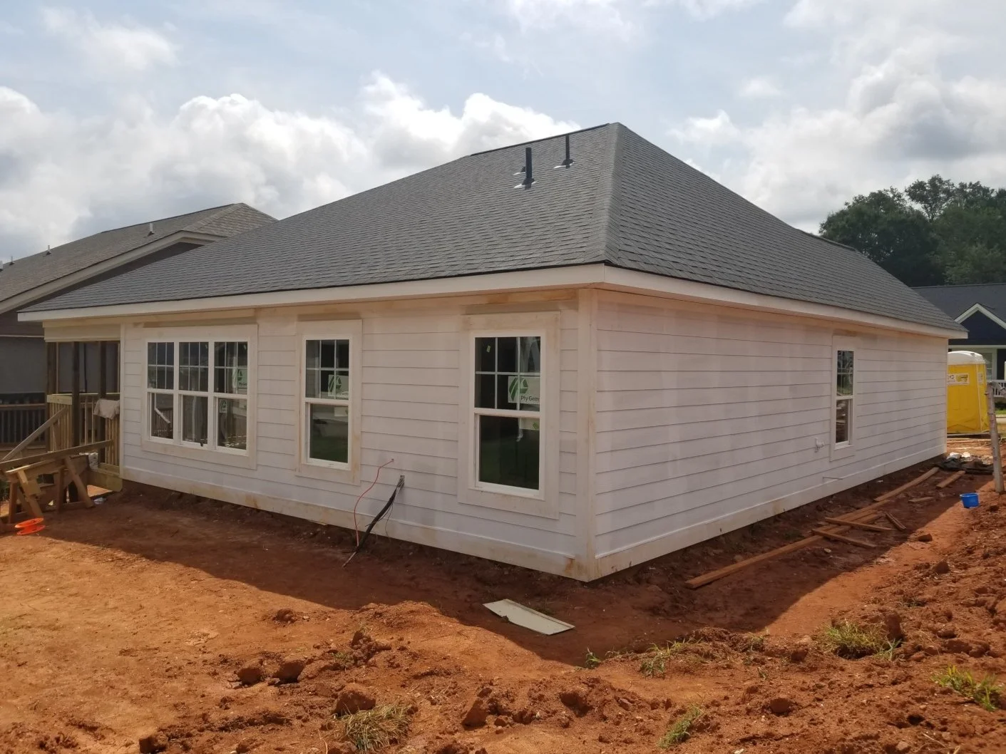 Newly constructed single-story house with white siding and a dark gray shingled roof, under construction, with exposed dirt yard and some construction materials around.