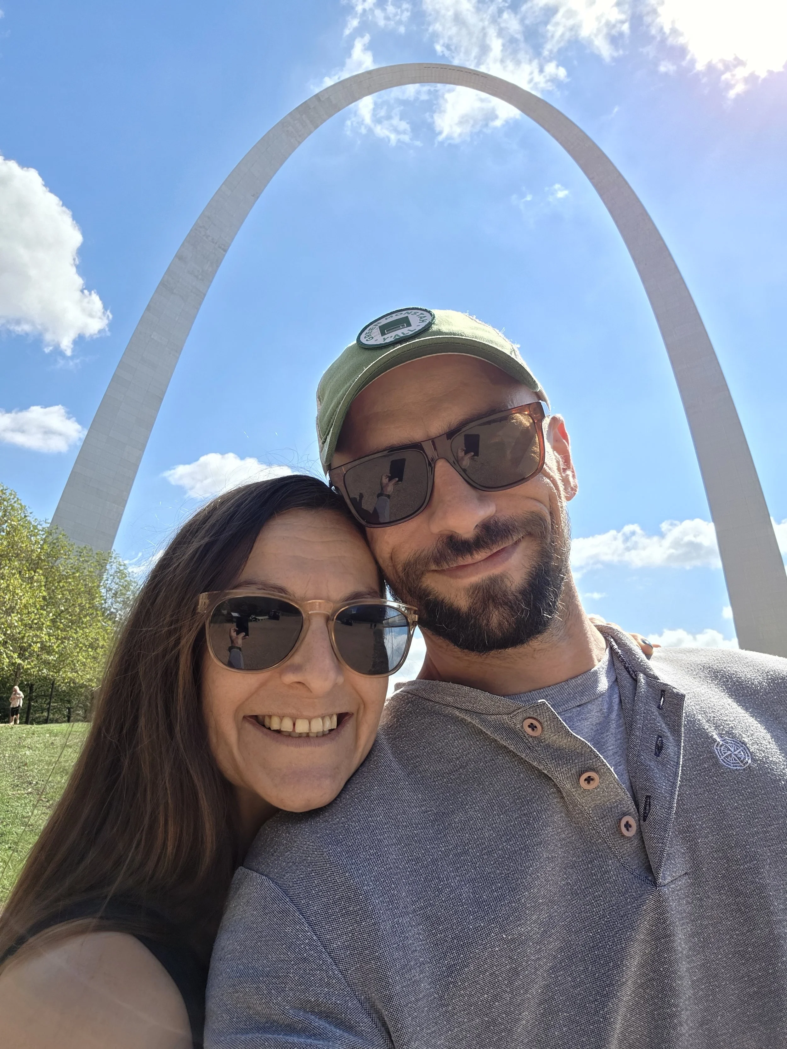 A smiling couple taking a selfie in front of the Gateway Arch in St. Louis on a sunny day. The woman has long brown hair and sunglasses, and the man has a beard, sunglasses, and a green cap.