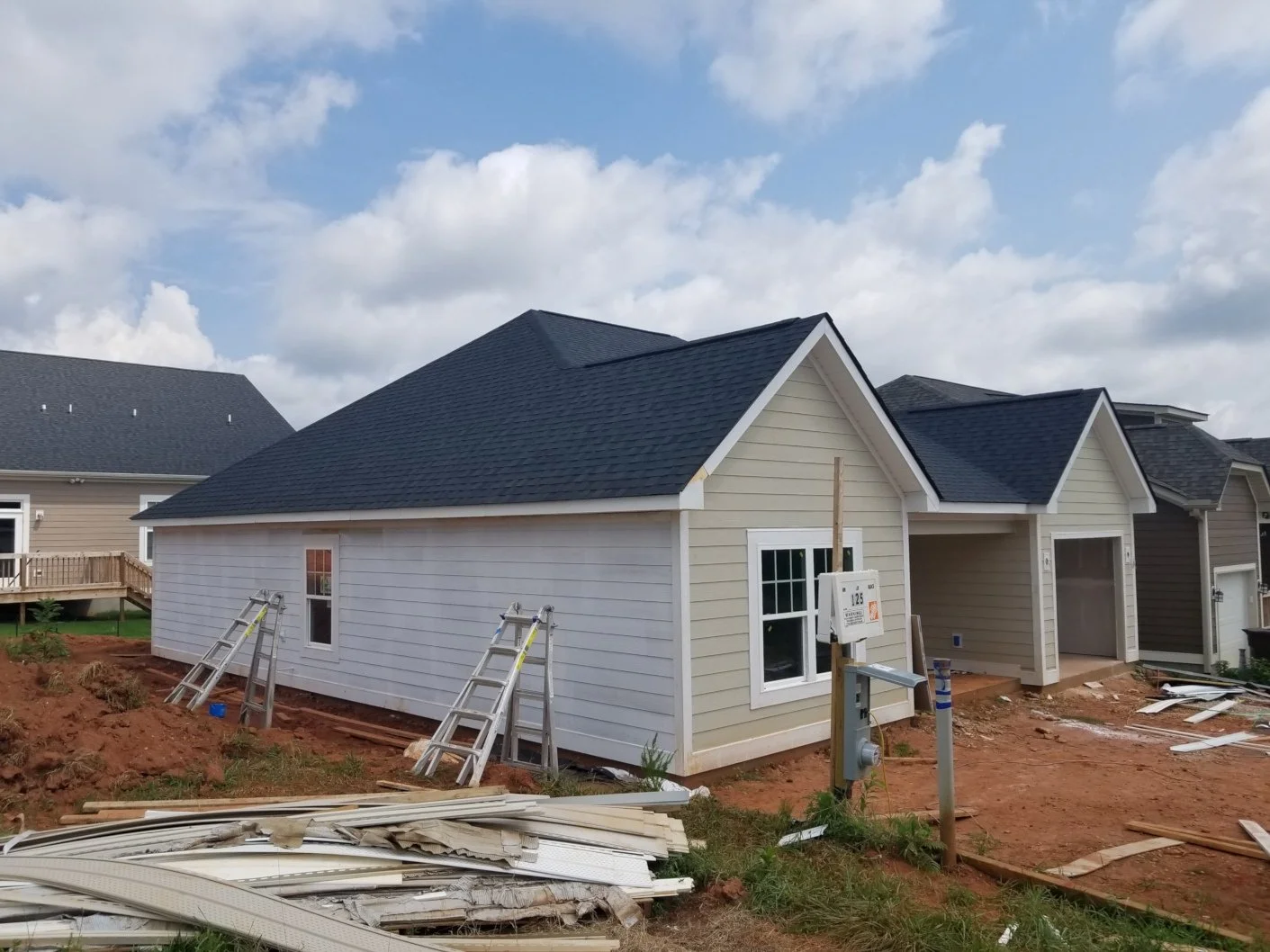 Under construction house with white siding, black roof, two windows, surrounded by construction debris and ladders, in a residential area with neighboring houses, blue sky, and clouds.