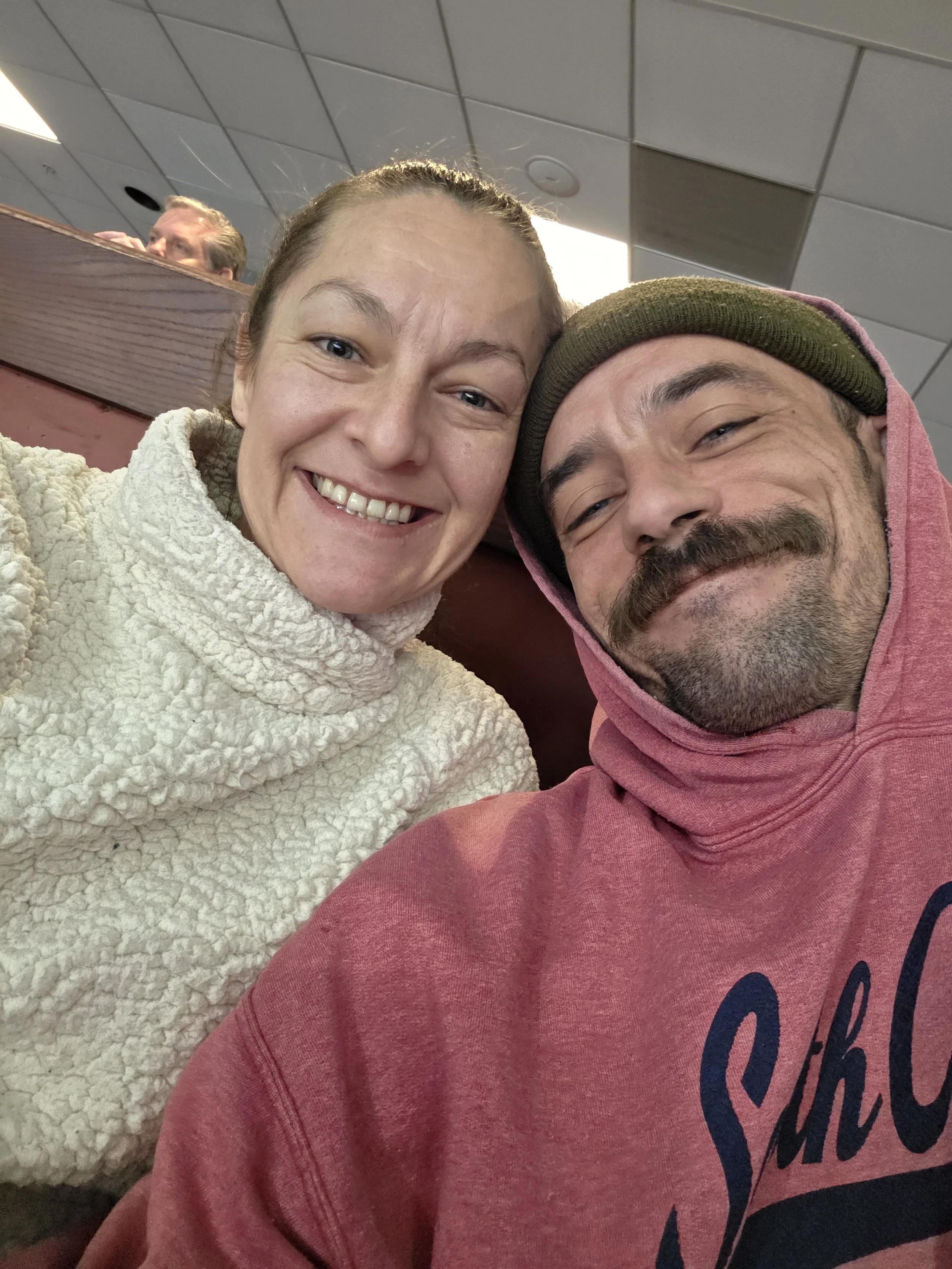 A woman and a man smiling and taking a selfie together inside an indoor public place, with a person visible in the background seating behind a red booth.