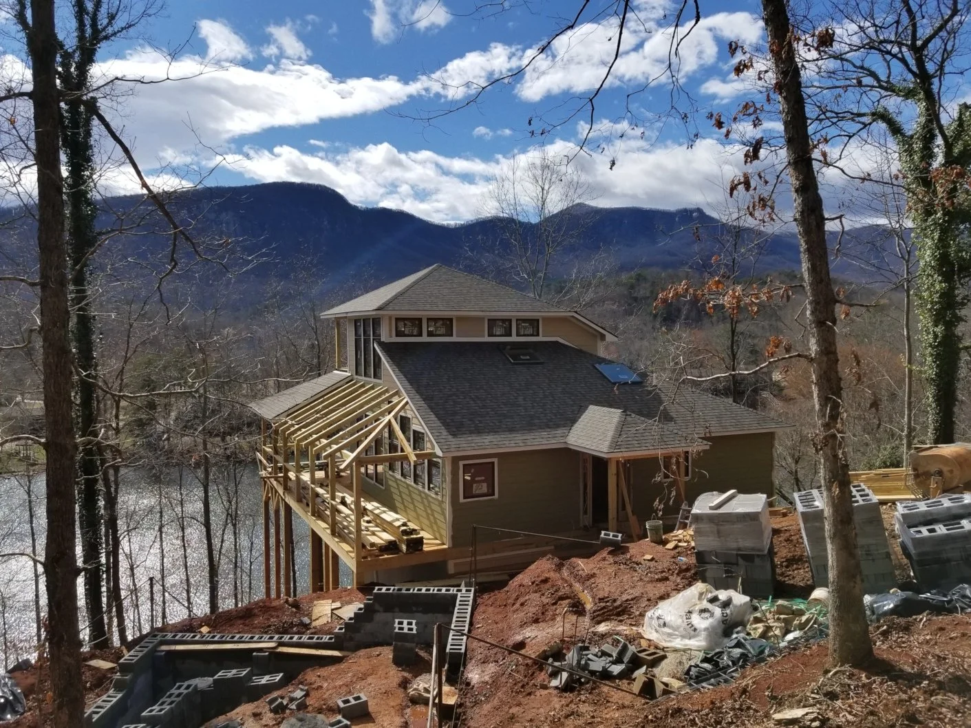 A house under construction on a hillside with a view of mountains and a river, featuring a partially built wooden deck, scattered building materials, and trees with autumn leaves.