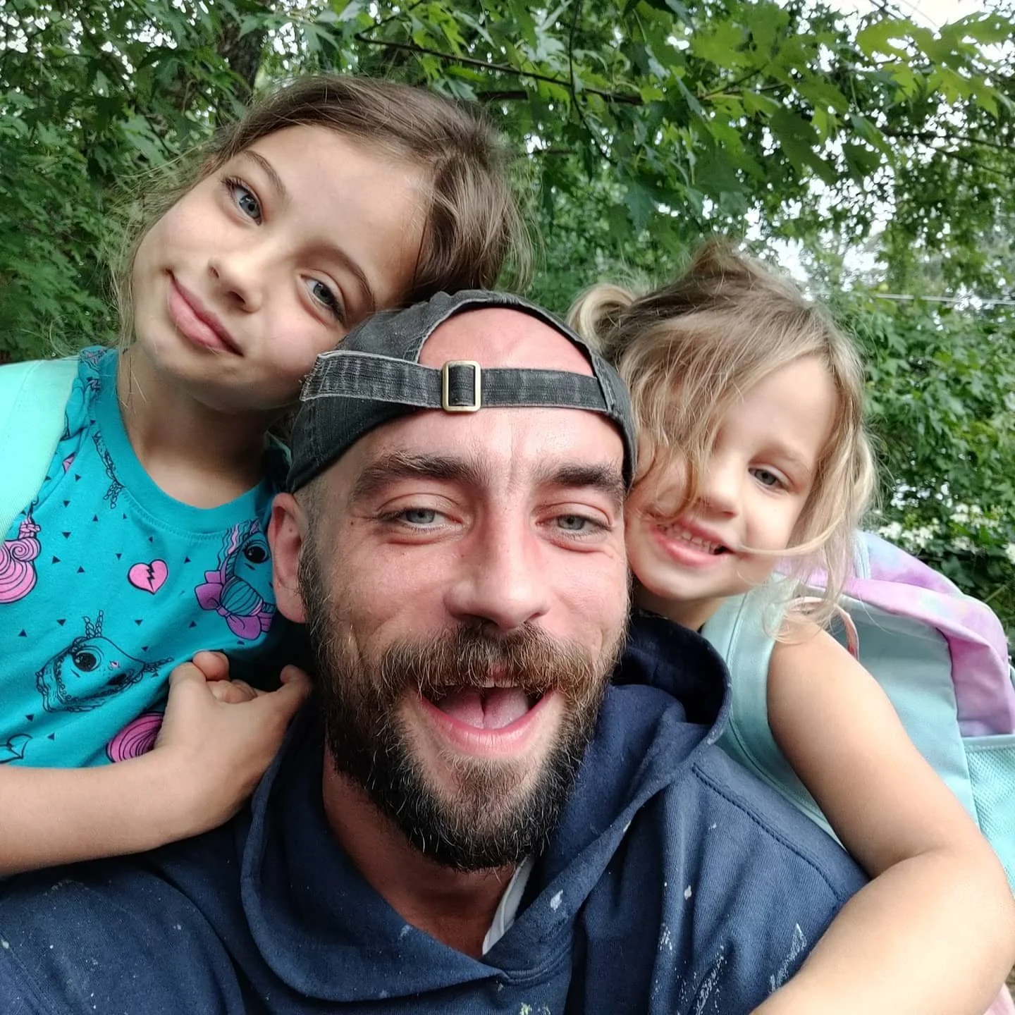 A man with a beard wearing a cap and hoodie is taking a selfie with two young girls, one on each side, outdoors with green trees in the background.