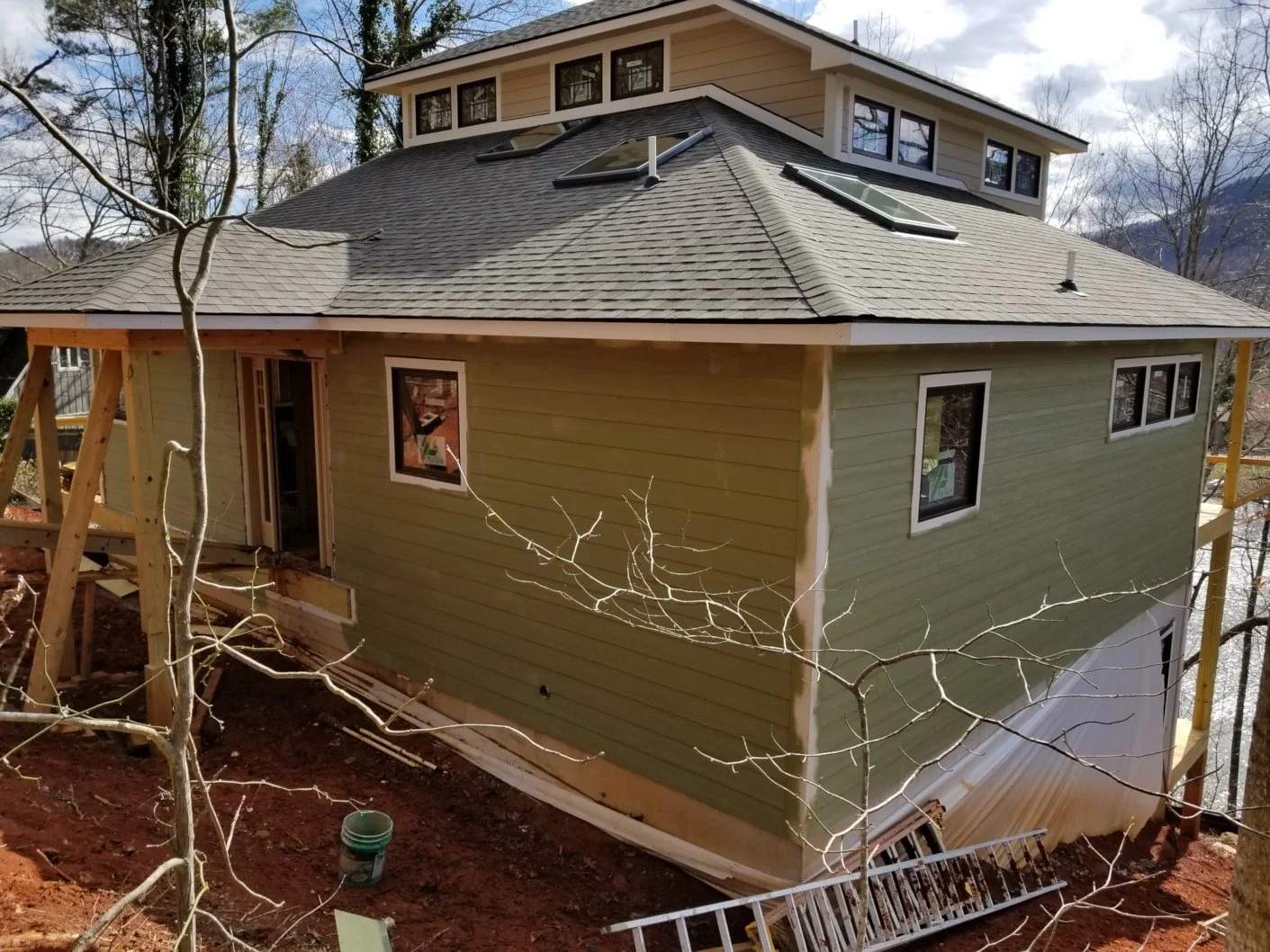 A house under construction with green siding, multiple windows, a multi-level roof with skylights, and wooden scaffolding around it, situated on a hillside with trees and mountains in the background.