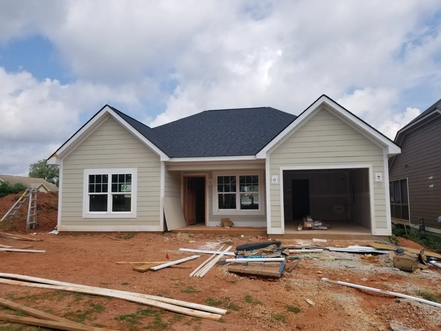 A house under construction with beige siding, white trim, and a dark shingle roof, featuring a two-car garage, with construction materials and tools scattered on the dirt yard in front.
