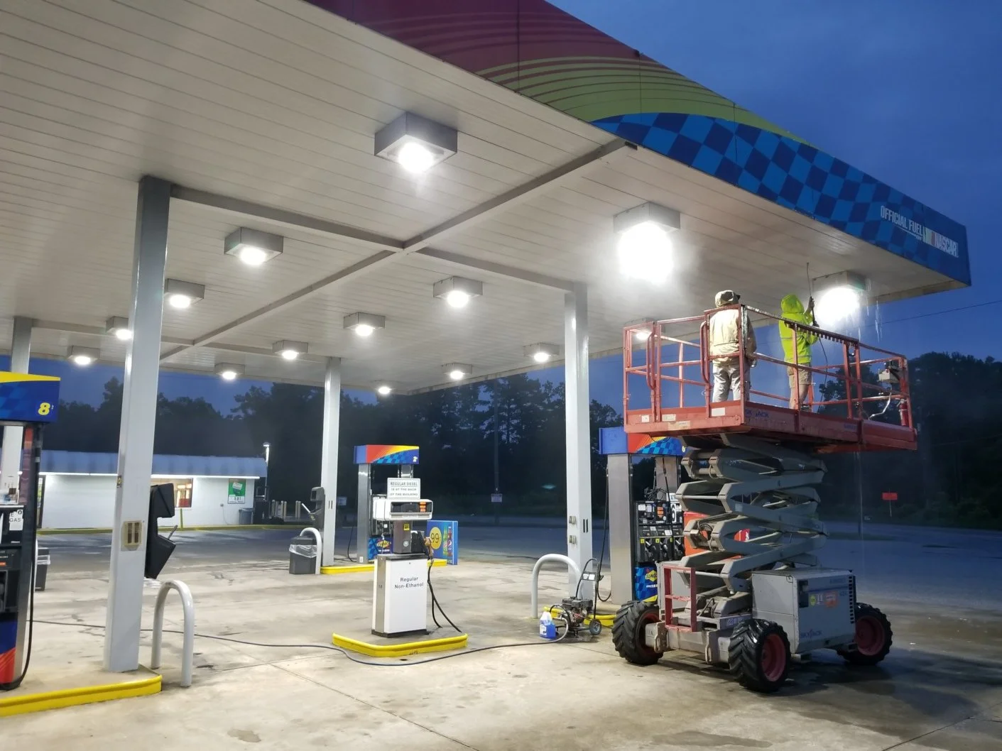 Two workers on a lift cleaning the ceiling of a gas station canopy at dusk. The gas station is well-lit and has multiple pumps.