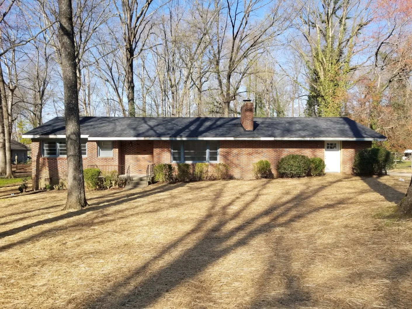 A single-story brick house with a black shingled roof, situated on a large yard with grass and trees, and a white door on the far right side.