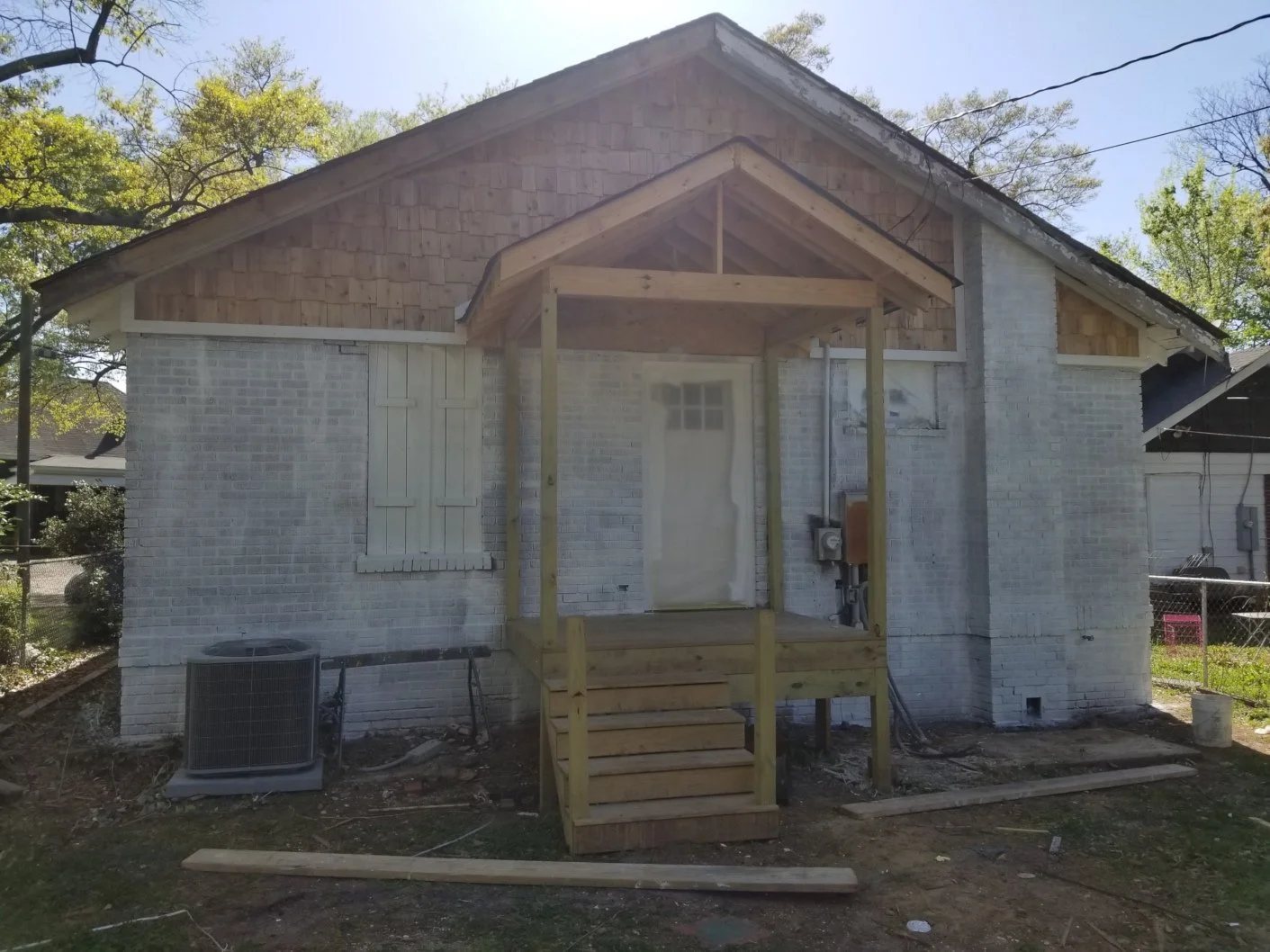Backyard view of a house under renovation with a new porch with steps and roof frame, white brick exterior, an air conditioning unit, and surrounding trees.