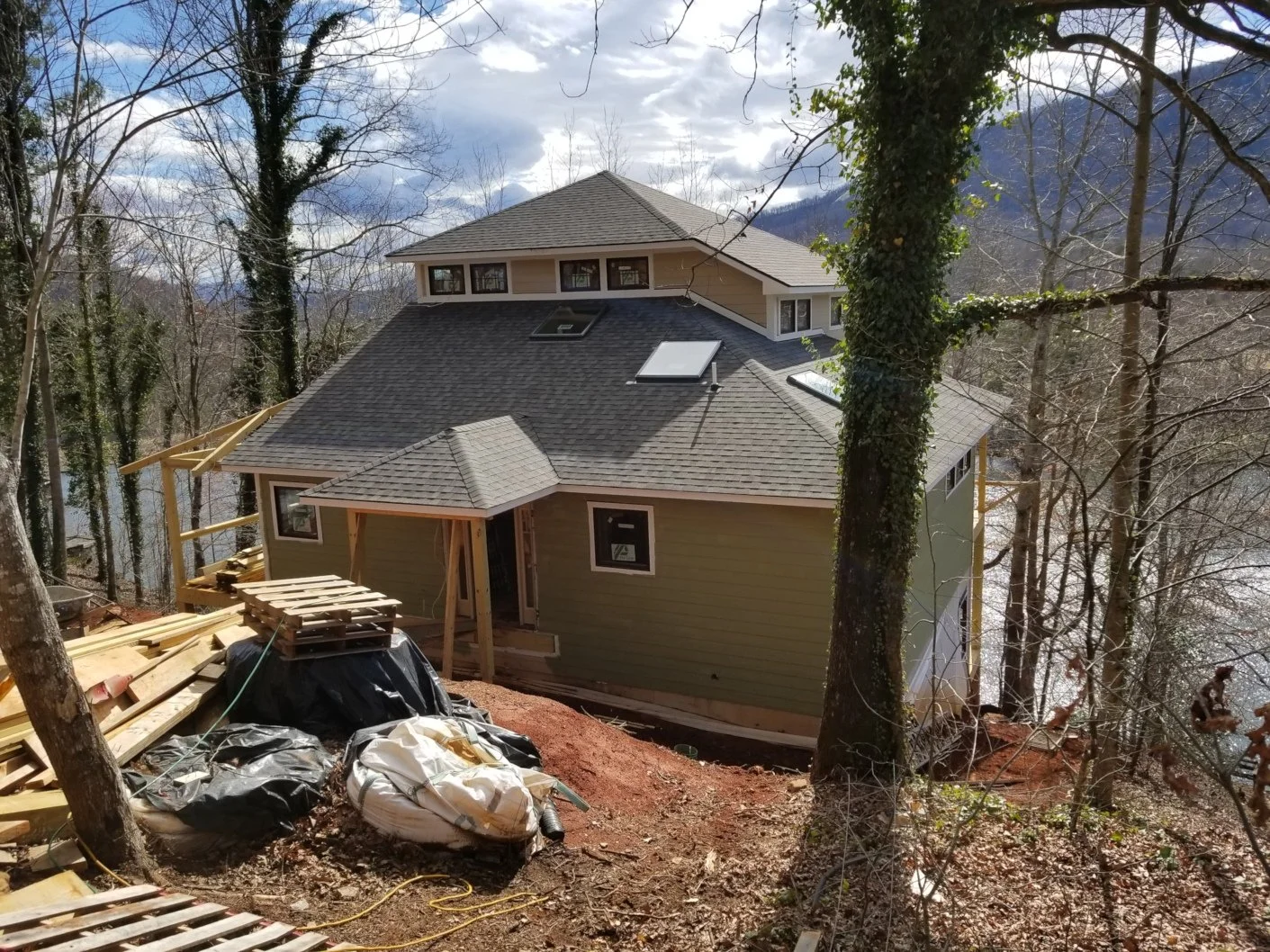 A house under construction on a wooded hillside, with scaffolding and construction materials in the foreground, trees around, and mountains in the background.