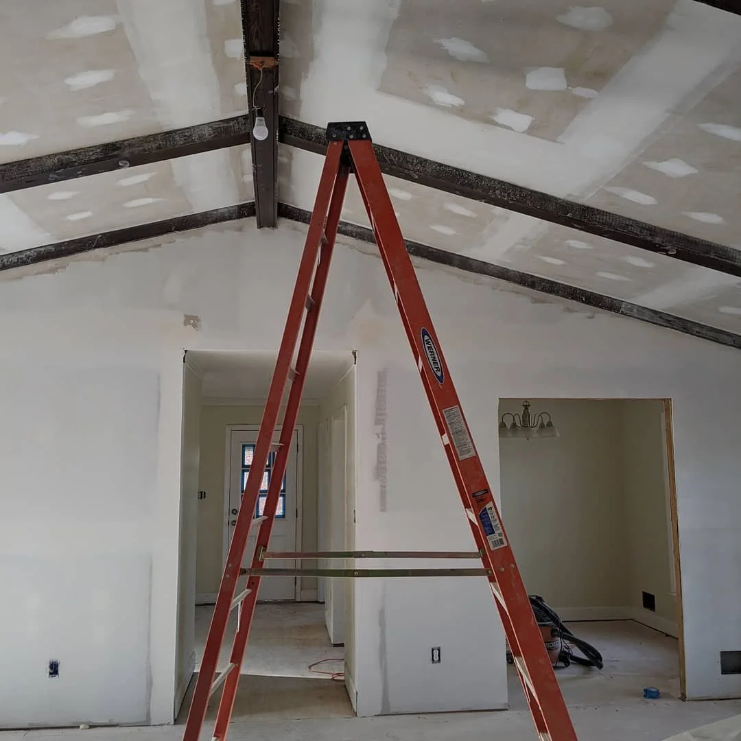 Interior of a room under construction with drywall walls, a partially finished ceiling, a red ladder in the center, and construction tools on the floor.