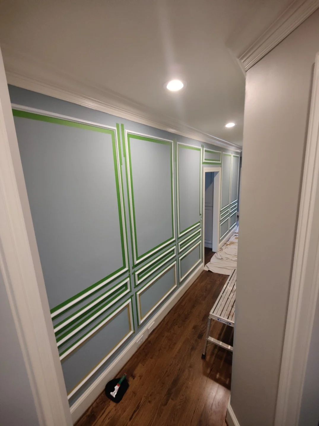 Hallway with light blue painted walls decorated with green and white geometric molding, hardwood floor, ceiling lights, and a small step ladder.