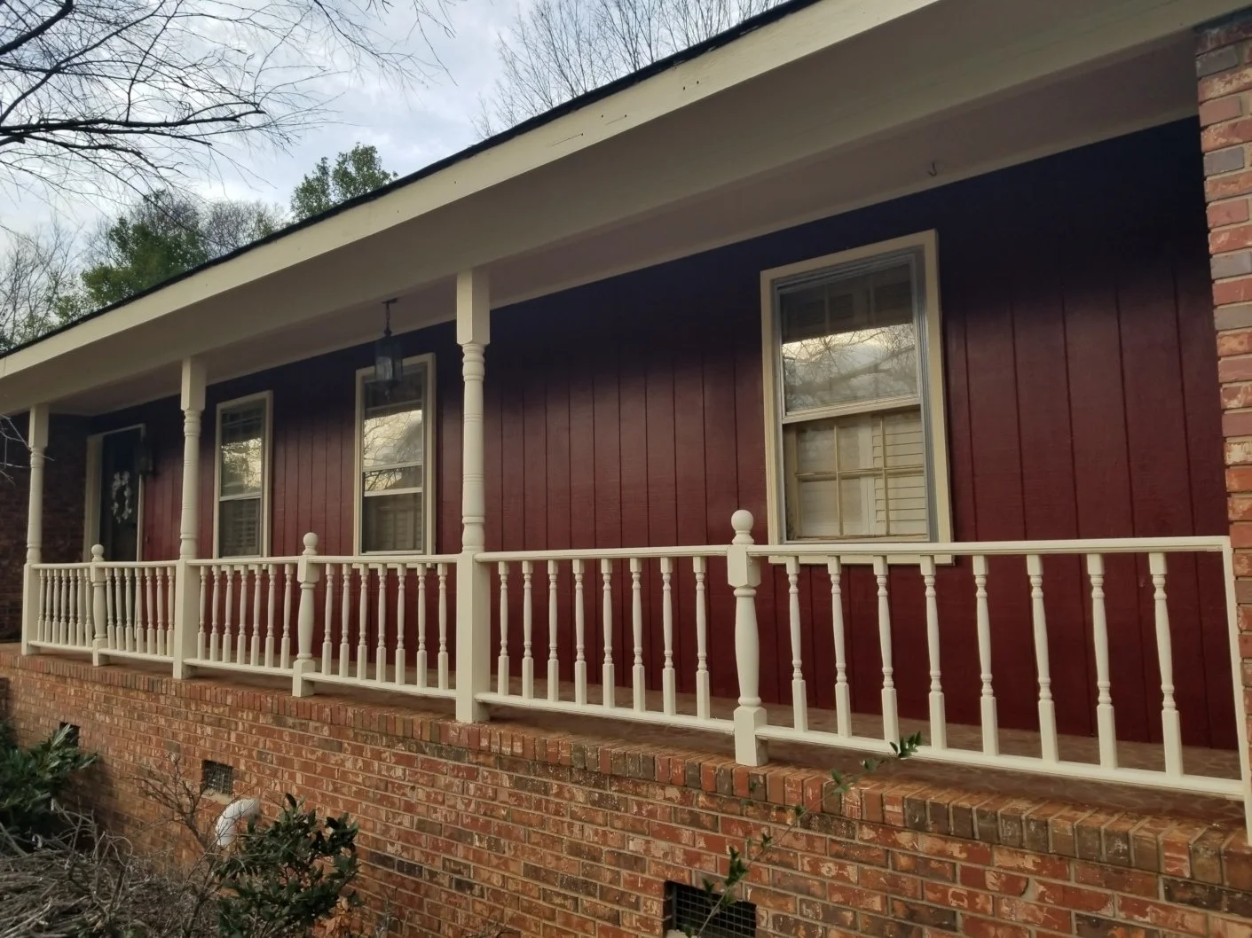 Front porch of a house with red wooden siding, white railing, brick foundation, and windows, surrounded by leafless trees.