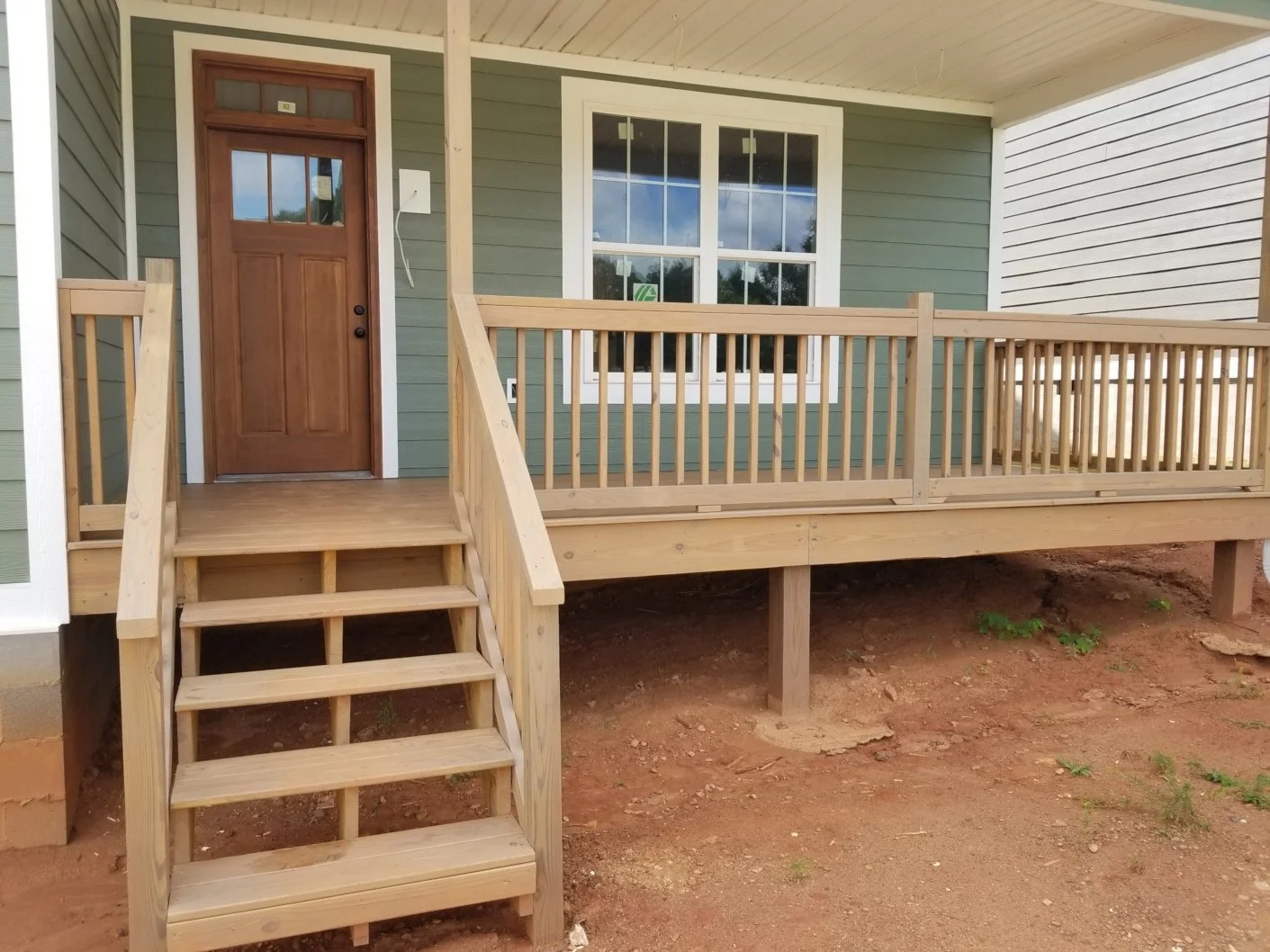 Newly constructed wooden front porch with stairs, door, and railing attached to a house with green siding and white trim
