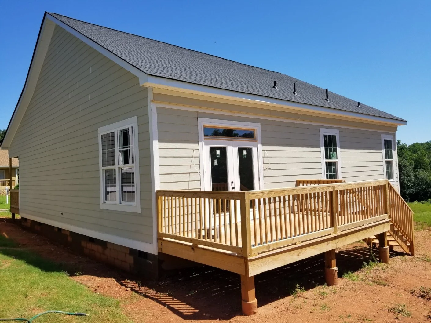 Newly built light-colored house with a wooden deck and stairs, surrounded by green grass and under a clear blue sky.