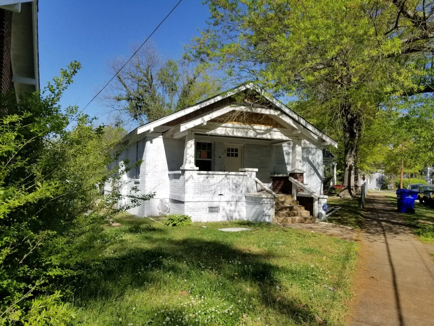 A white brick house with a small front porch and steps, surrounded by green trees and grass, on a sunny day with a clear blue sky.