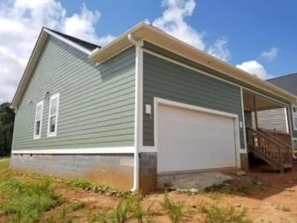 A house with green siding and a white garage door, located on a grassy lot under a partly cloudy sky.