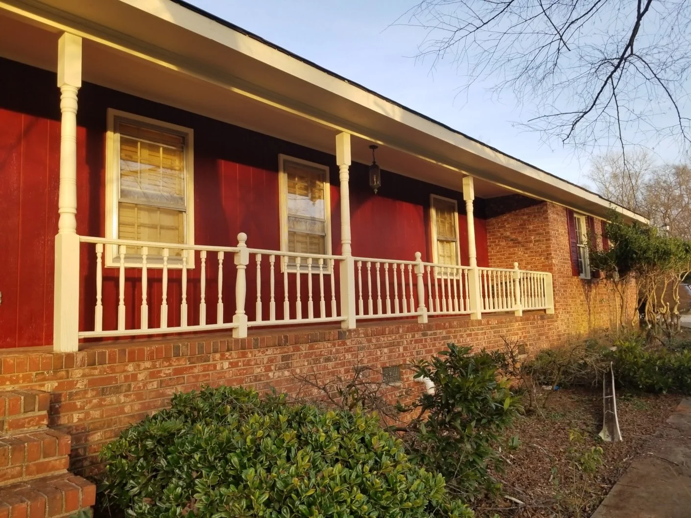 Front of a house with a brick foundation, red painted walls, white porch railing, and beige columns. There are three windows with wooden shutters, and a porch light hanging from the ceiling. Bushes and small plants are in the garden in front.