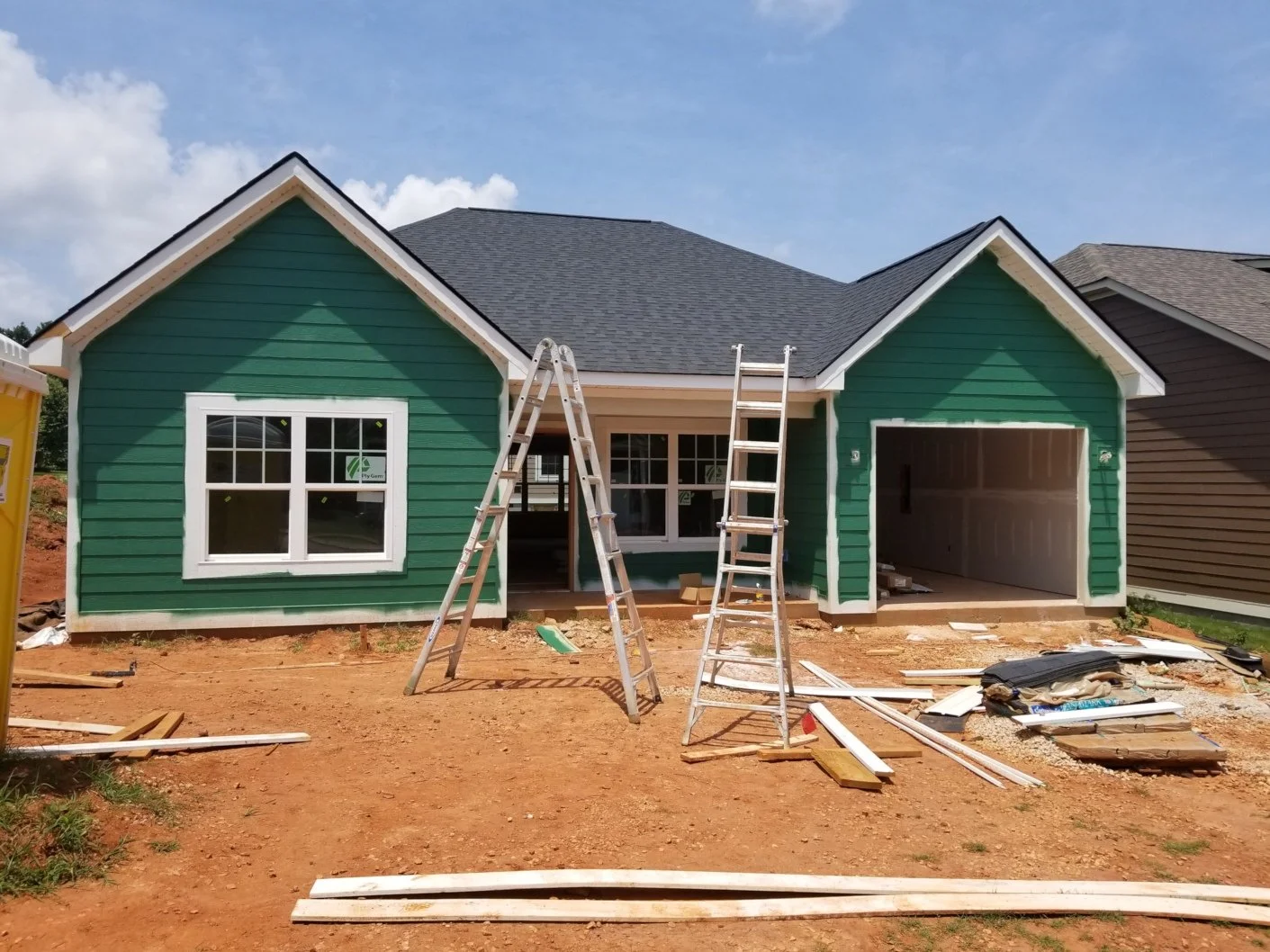 A house under construction with green siding, white trim, and a dark gray roof. Two ladders are positioned in front, and construction materials and tools are scattered on the reddish dirt yard.