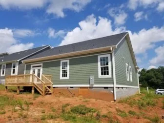 Newly constructed green house with white window frames and a small wooden deck, on a dirt lot with a partly cloudy sky.