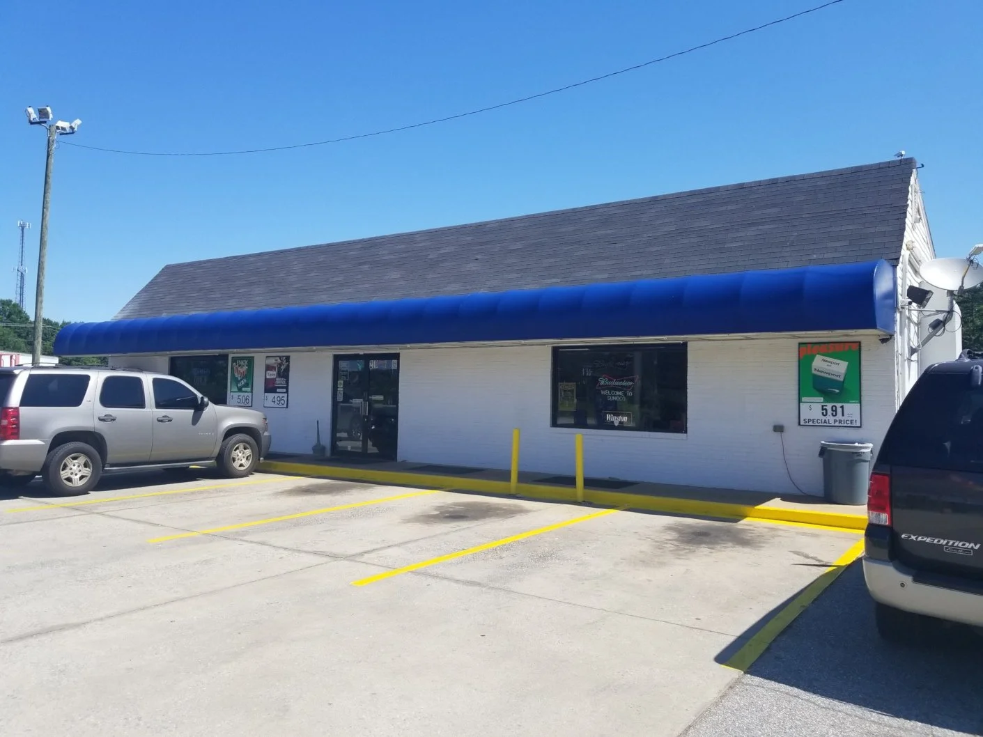 Single-story convenience store with a blue awning and parking lot in front, a gray SUV parked, and a black SUV partially visible on the right. Clear blue sky overhead.