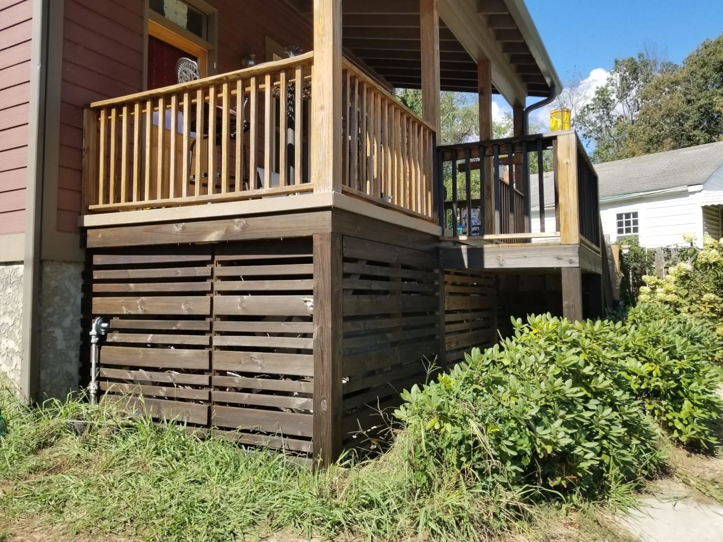 Photo of a wooden deck with railings attached to a house, with grass and bushes underneath, and neighboring houses in the background.