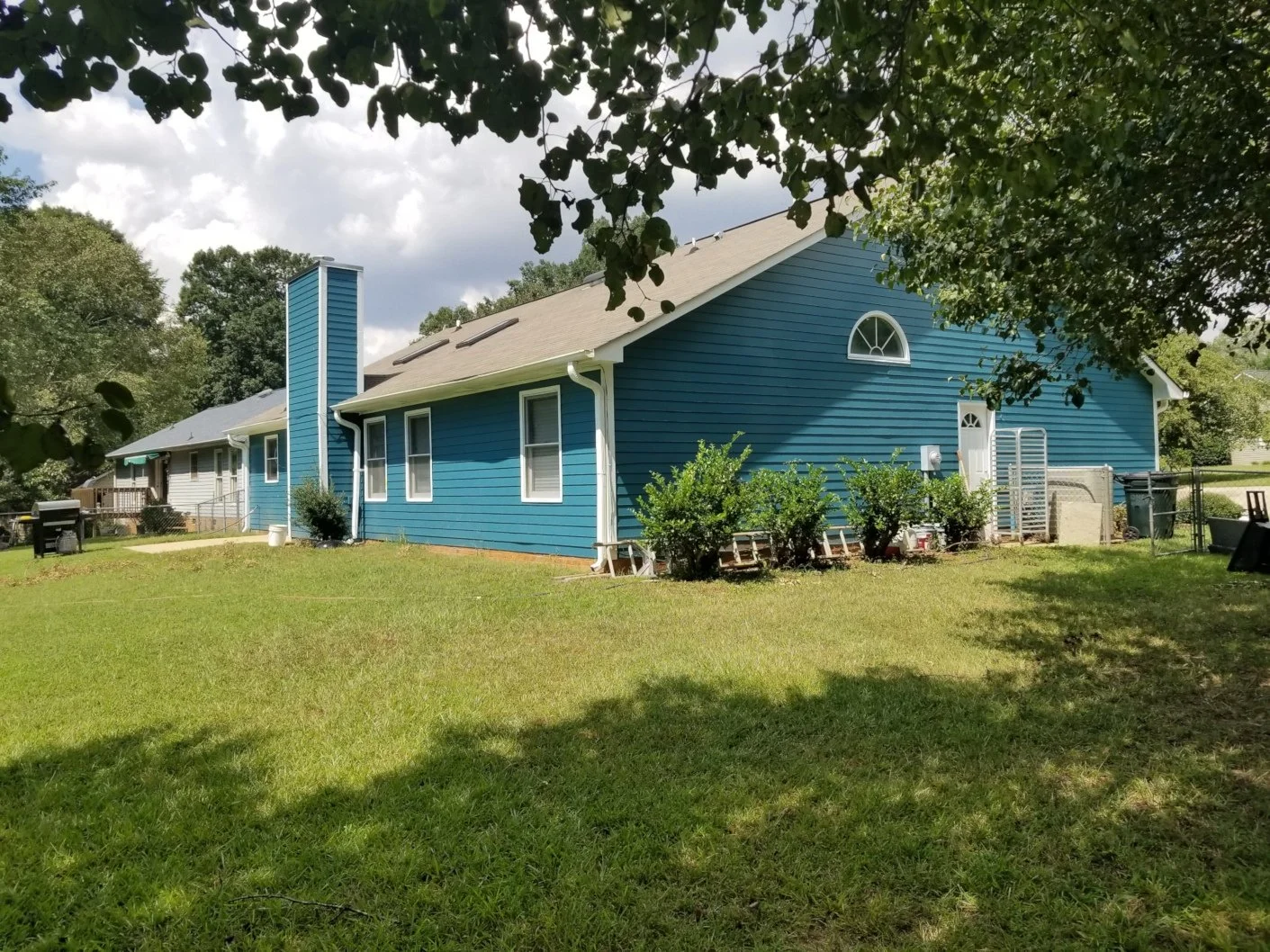 A blue house with white trim and a chimney, surrounded by a grassy yard and trees, under a partly cloudy sky.