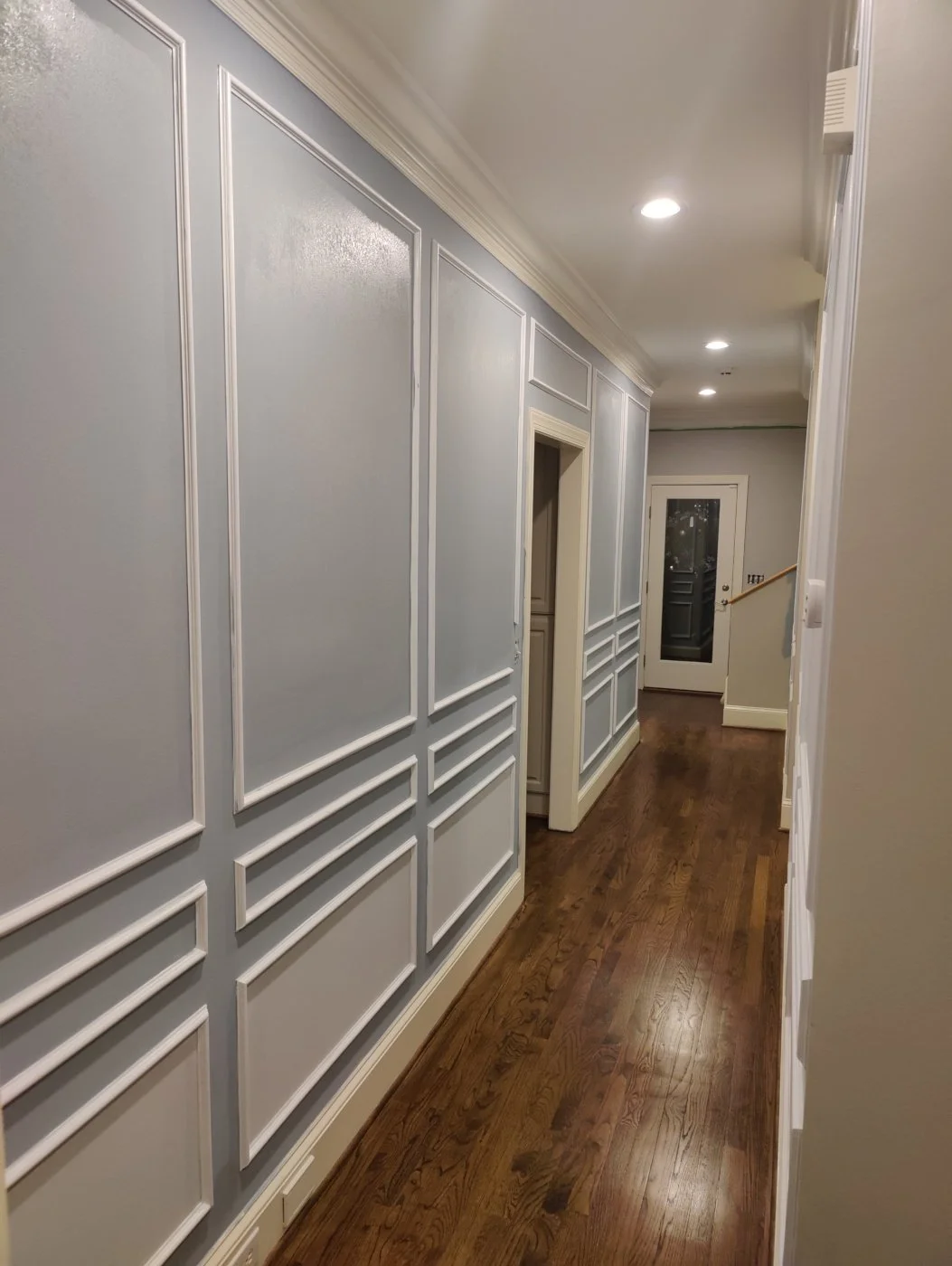 A hallway with light blue painted walls featuring decorative molding, hardwood floors, and recessed ceiling lights. At the end of the hallway, there is a door with a window.