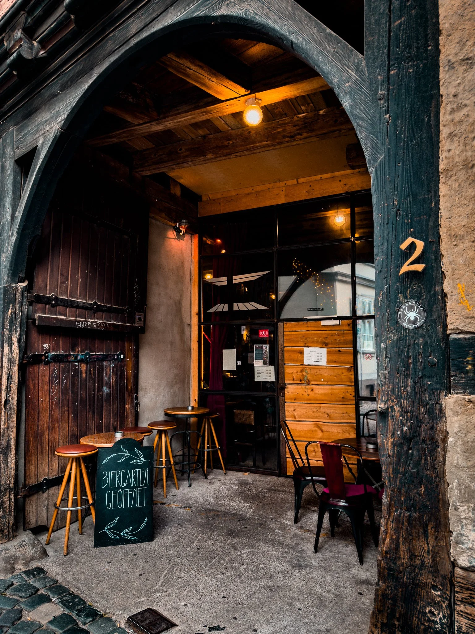 Eingang zu einem Biergarten mit einer Tafel, die 'Biergarten geöffnet' schreibt, und mehreren Stühlen davor.