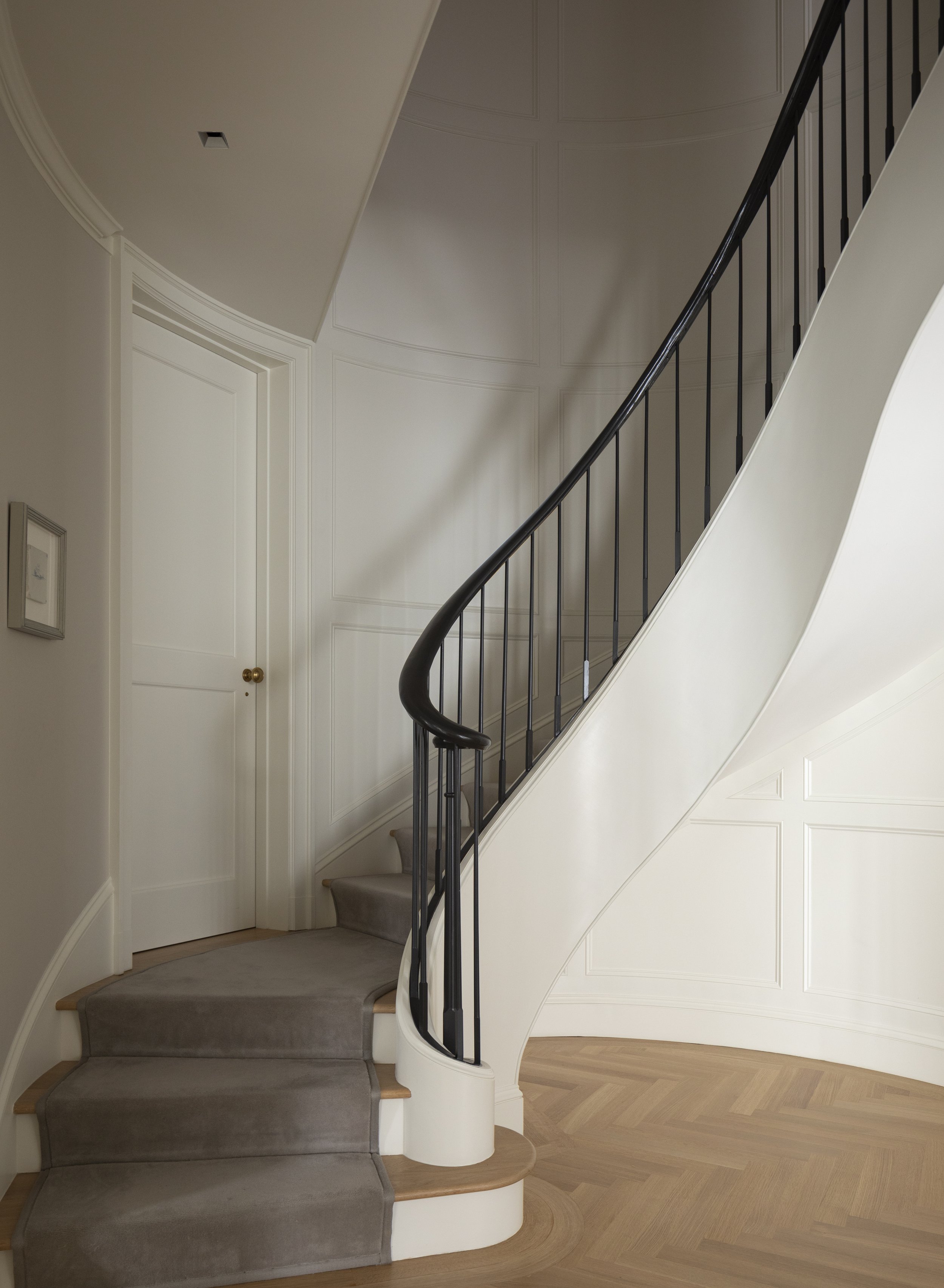 Interior of a house featuring a curved staircase with wooden steps, black metal railing, and a beige carpet runner, adjacent to a white wall with decorative paneling and a closed white door with a brass doorknob.