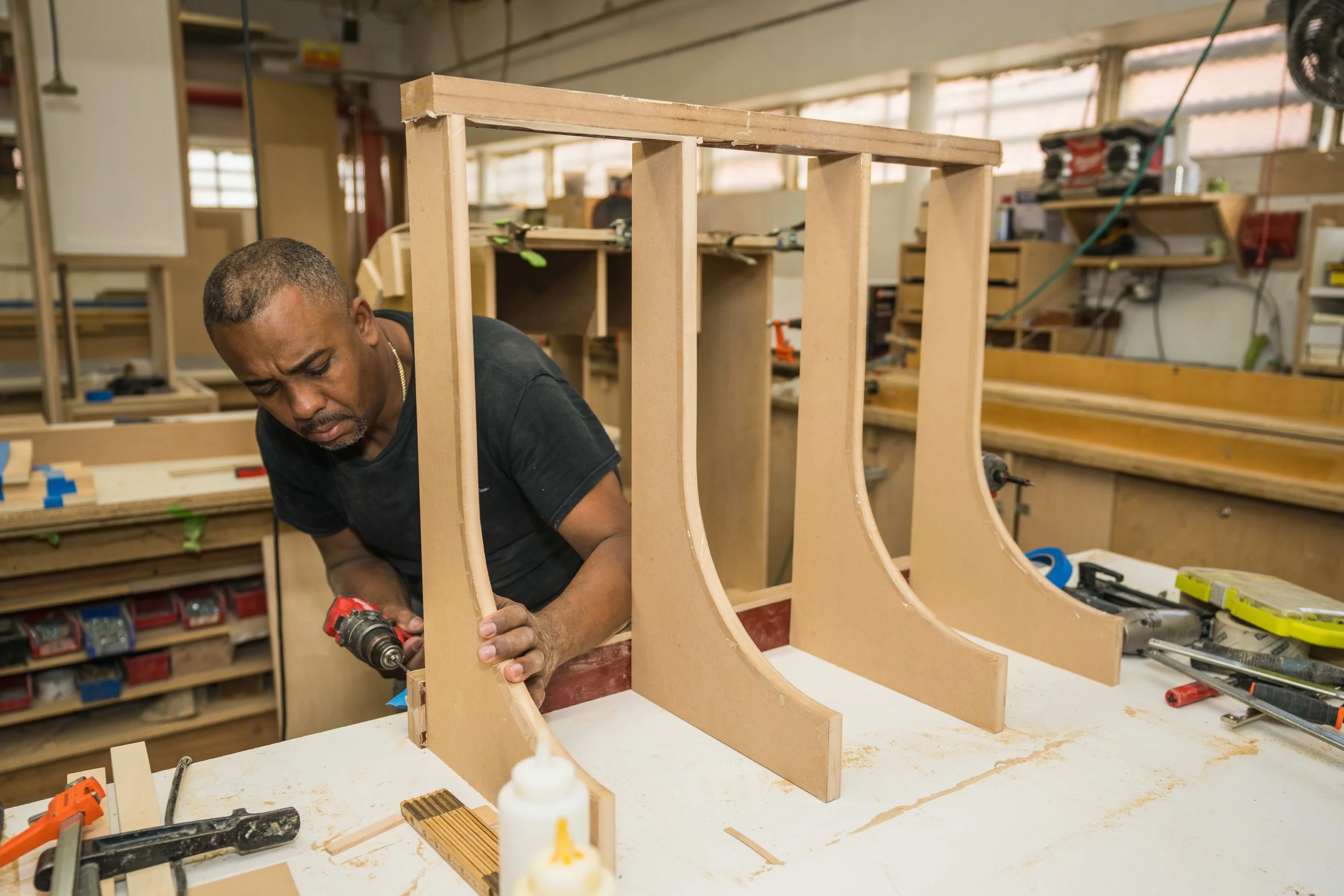 A man working on assembling a wooden structure in a woodworking shop, with various tools and materials around him.