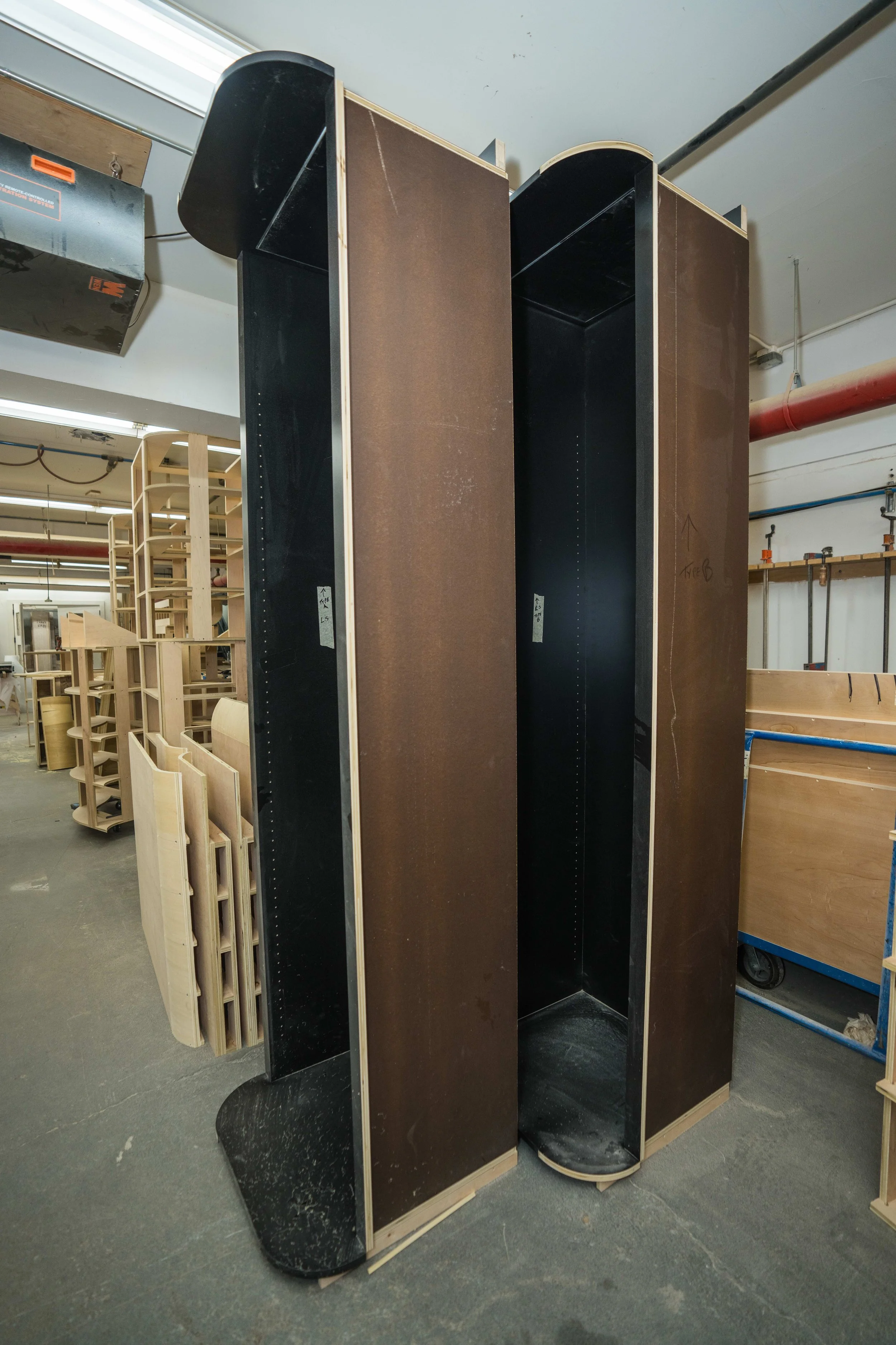 Two tall, black, oval-shaped display shelves with wooden edges in a workshop or storage area, with some wooden furniture and equipment in the background.