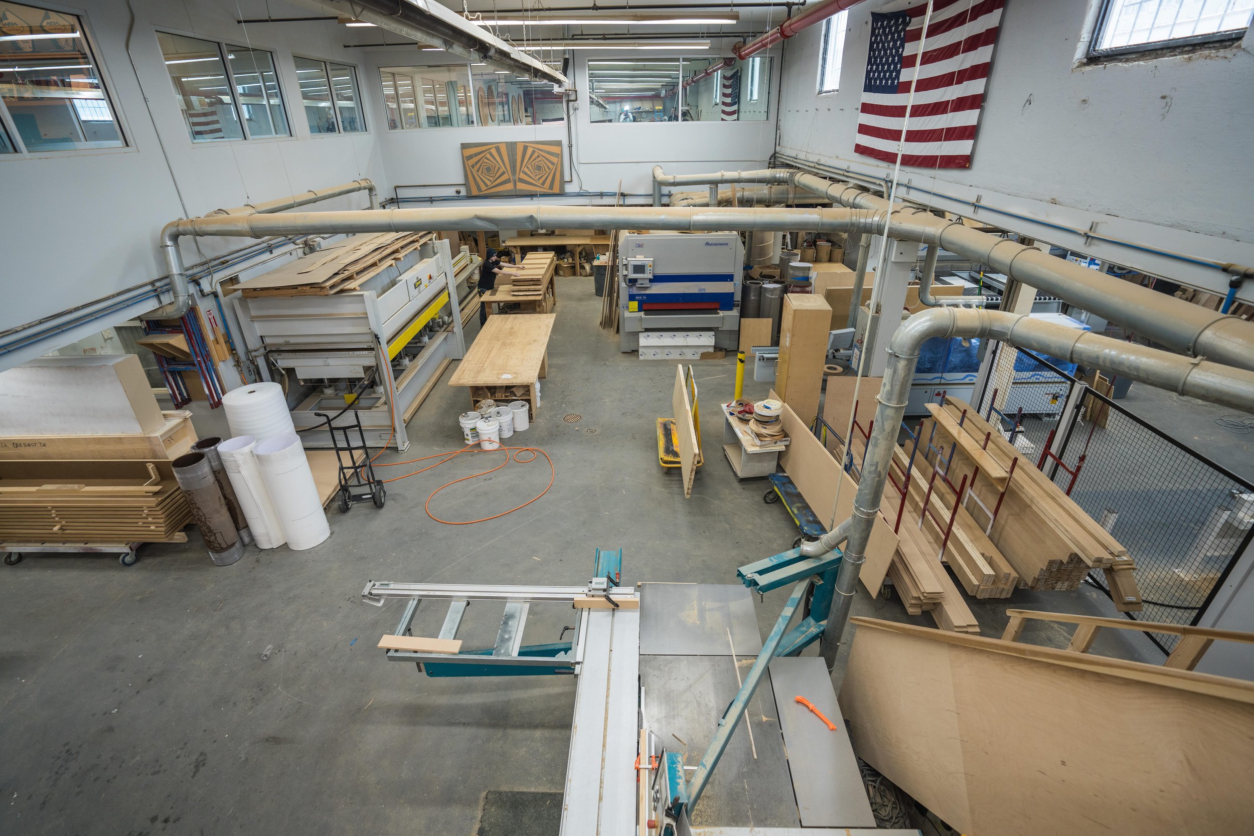 Woodworking workshop with various tables, machinery, and stacks of wood; American flag hanging on the wall.