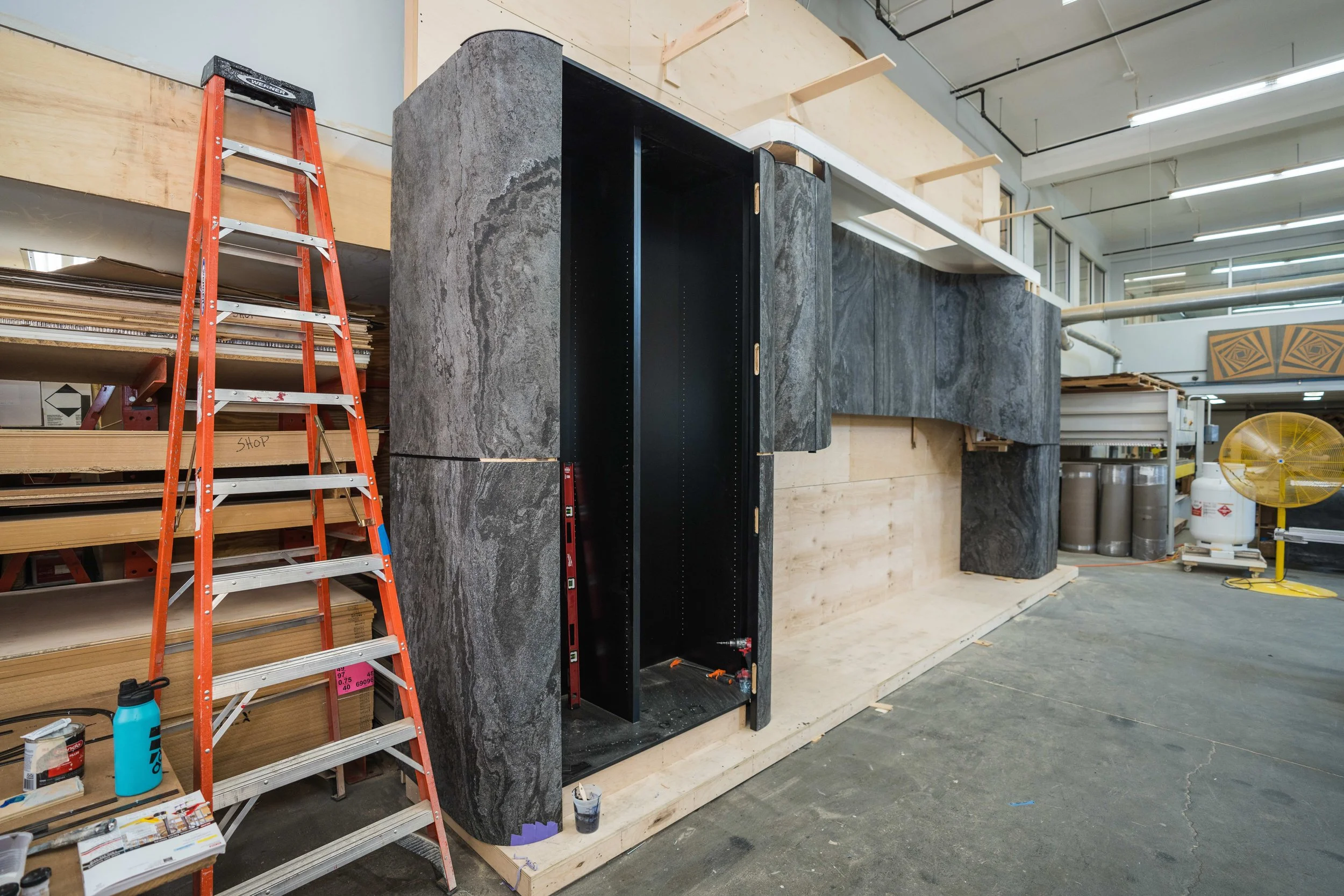 Construction site with a wooden wall behind which is a large black and gray stone structure, a red ladder, shelves with wood, and construction tools.
