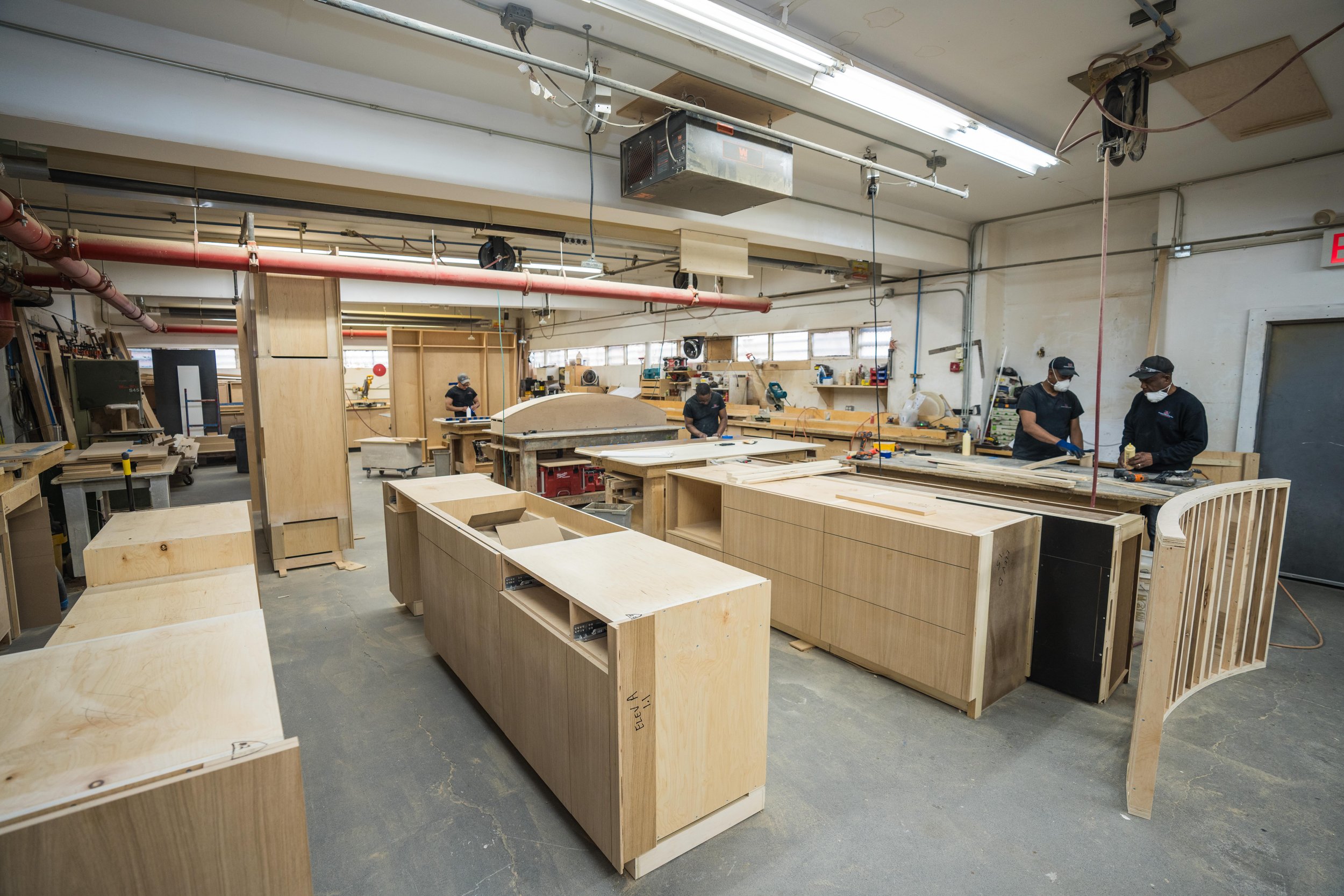 Woodworking workshop with workers assembling furniture, using tools and large wooden panels, in a well-lit industrial space.