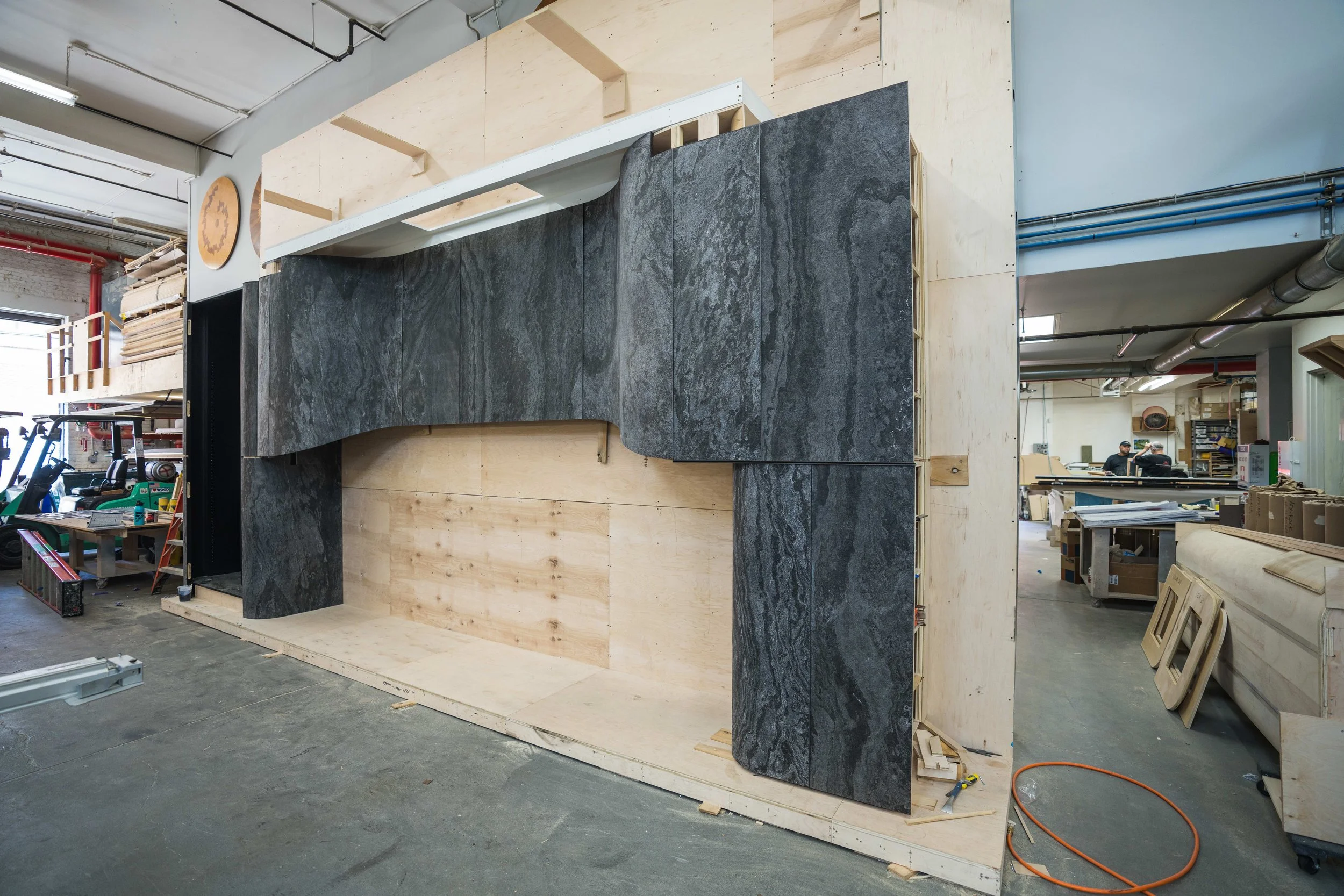 Interior of a workshop or showroom with a large wall structure made of wood and dark gray stone slabs. The wall appears to be under construction or installation, with tools and materials around. In the background, there are two people working and var