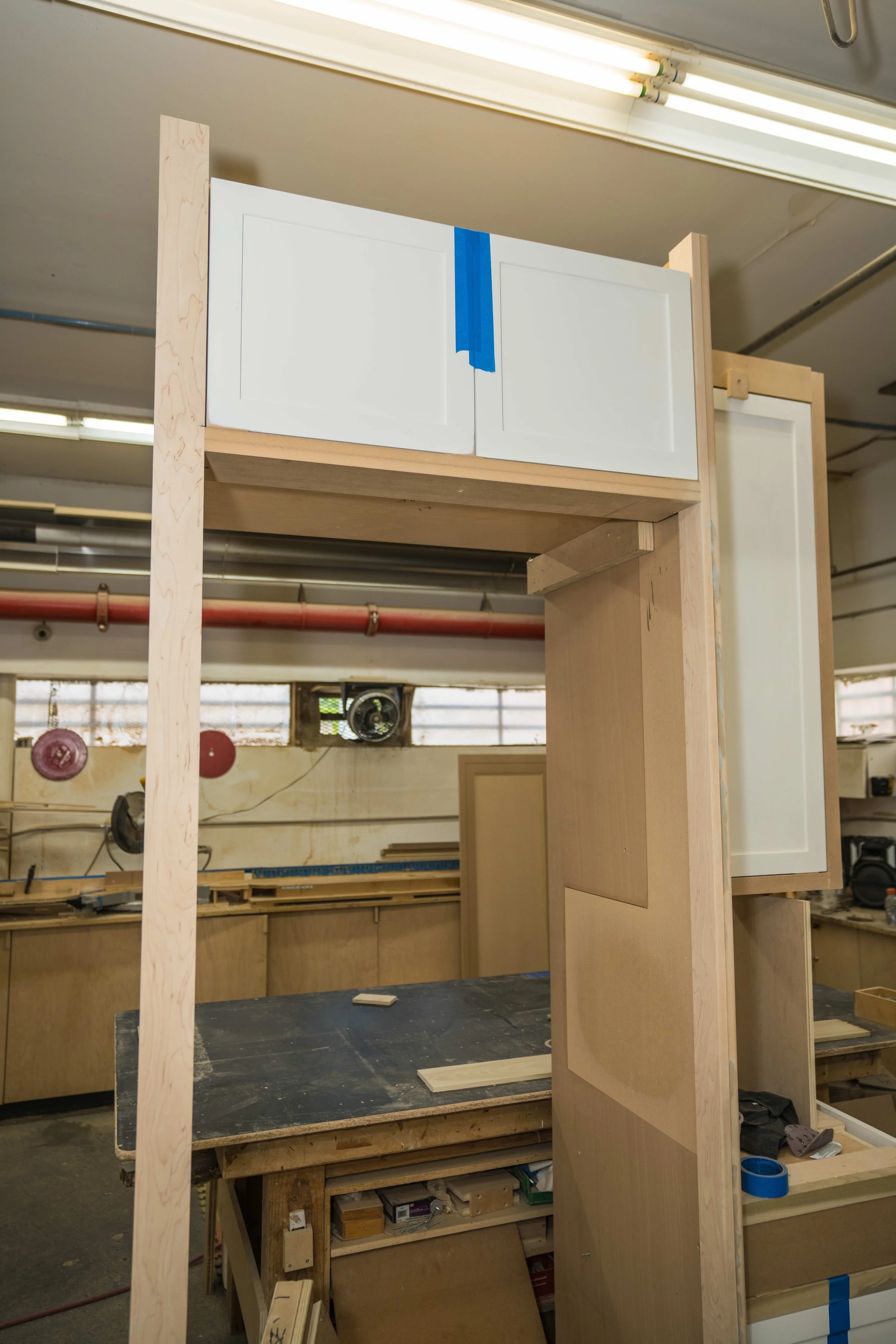 Woodwork in progress in a workshop with cabinets or furniture frames being assembled. Wooden panels and workspace tools are visible.