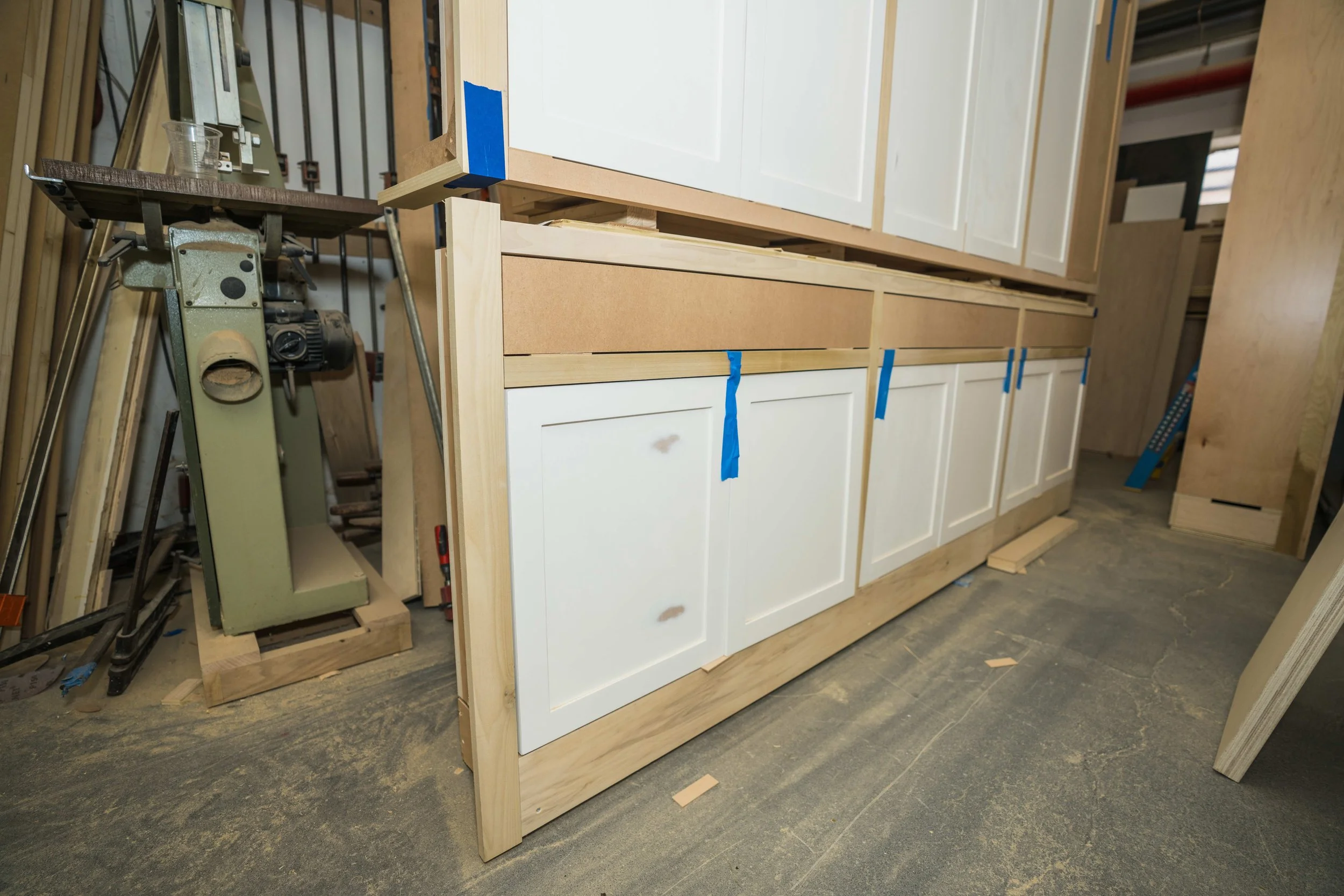 Wooden cabinet frame under construction in a workshop, with white cabinet doors installed and blue painter's tape on some edges.