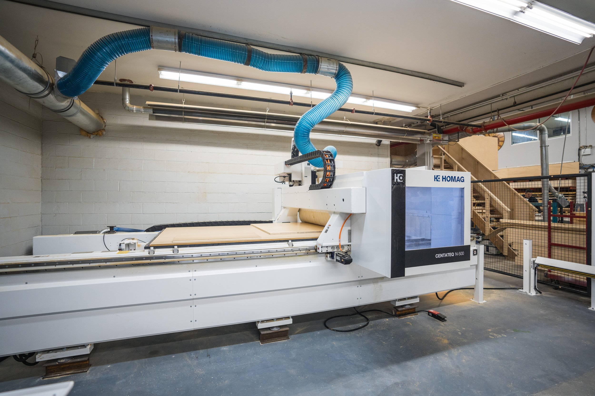 Large CNC woodworking machine in a workshop, with blue ventilation hoses and a staircase in the background.