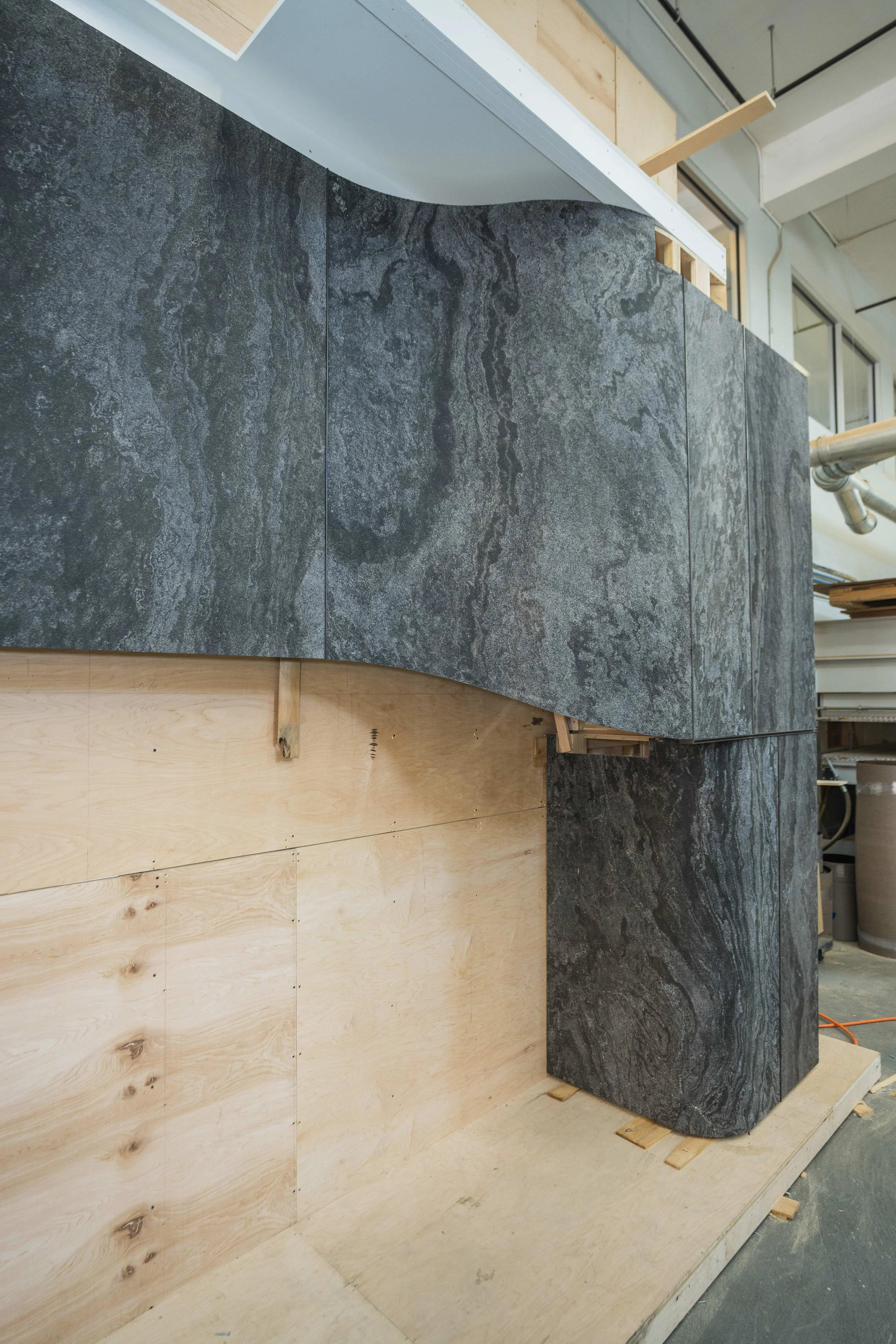 Interior under construction showing a curved wall with black stone tiles and a lower section of unfinished light wood paneling. Visible ceiling pipes, windows, and construction materials.
