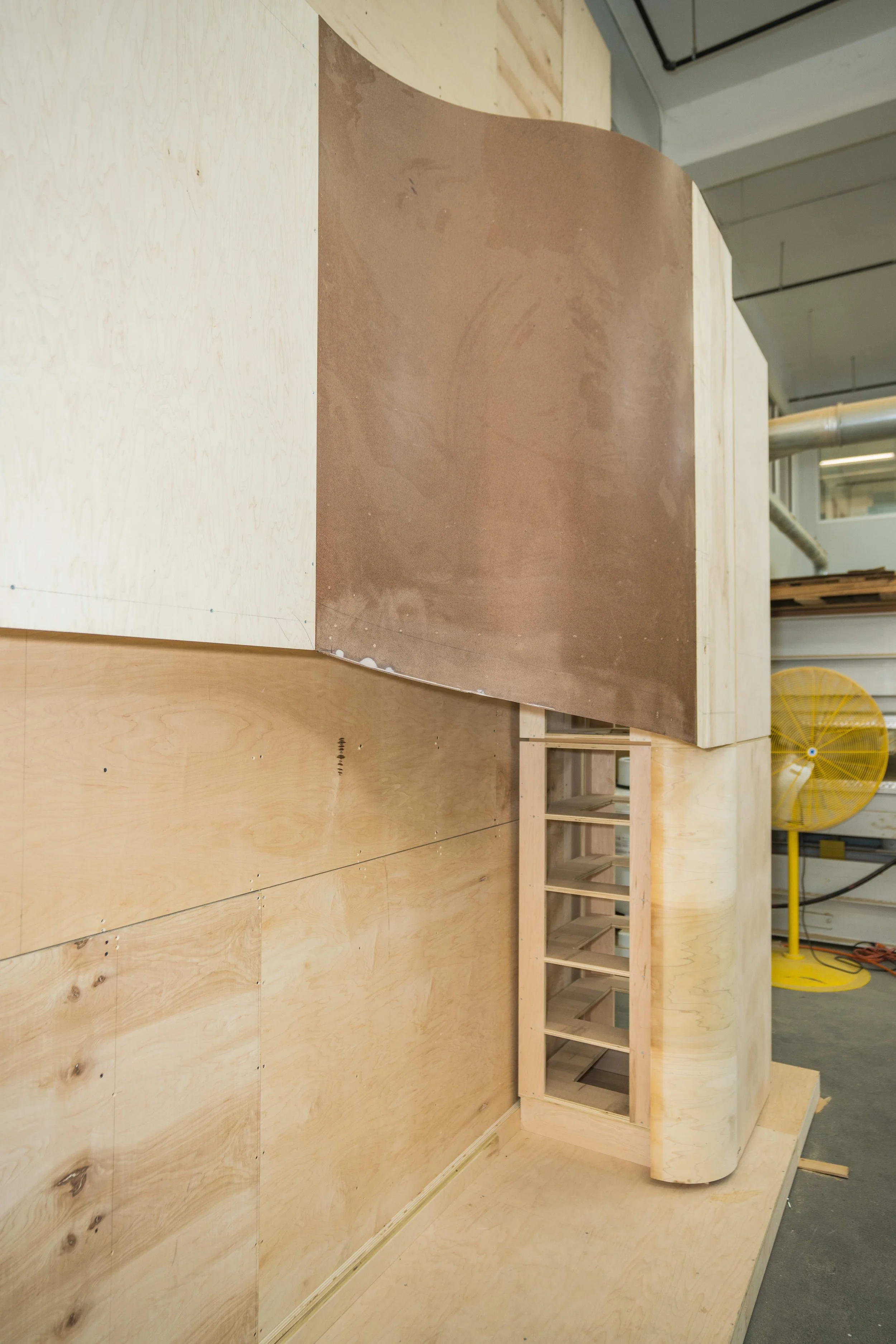 Wooden wall and structure under construction in a workshop or warehouse setting, with a yellow industrial fan in the background.